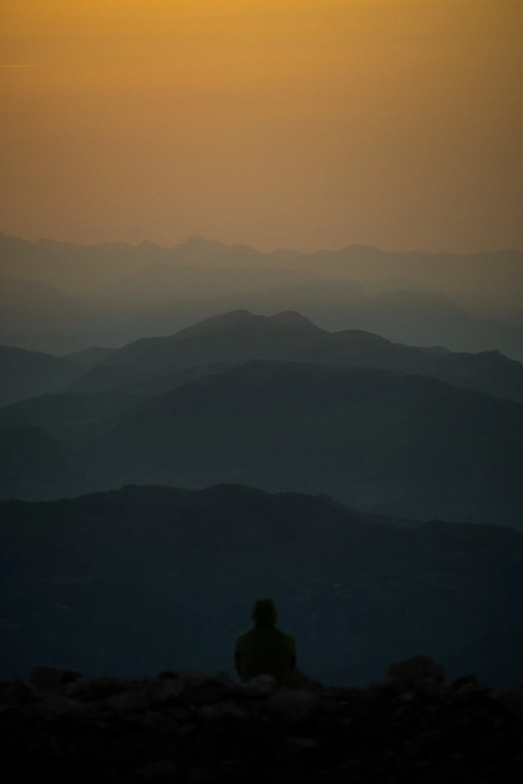 Silhouette of a seated person overlooking layered mountain ranges at sunset