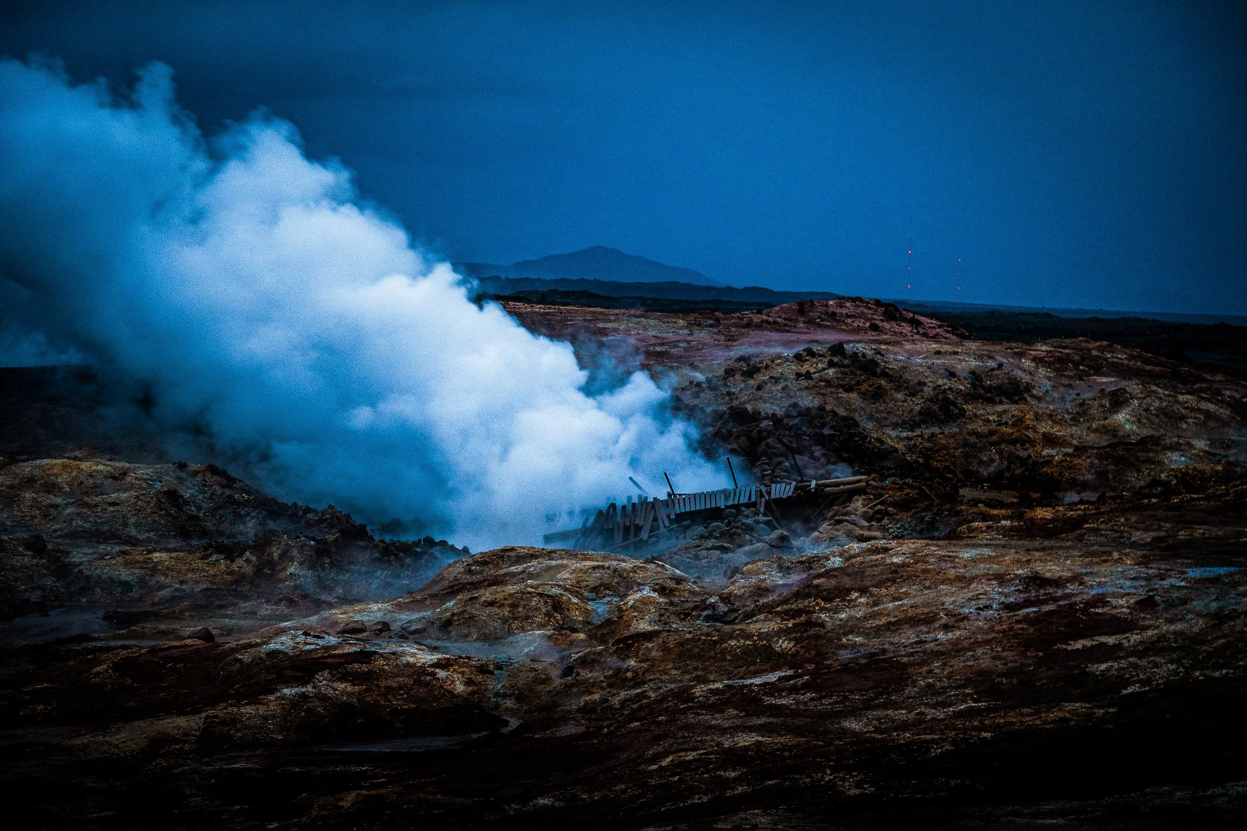Steaming geothermal hot spring surrounded by volcanic rocks, with mountains in the background at dusk