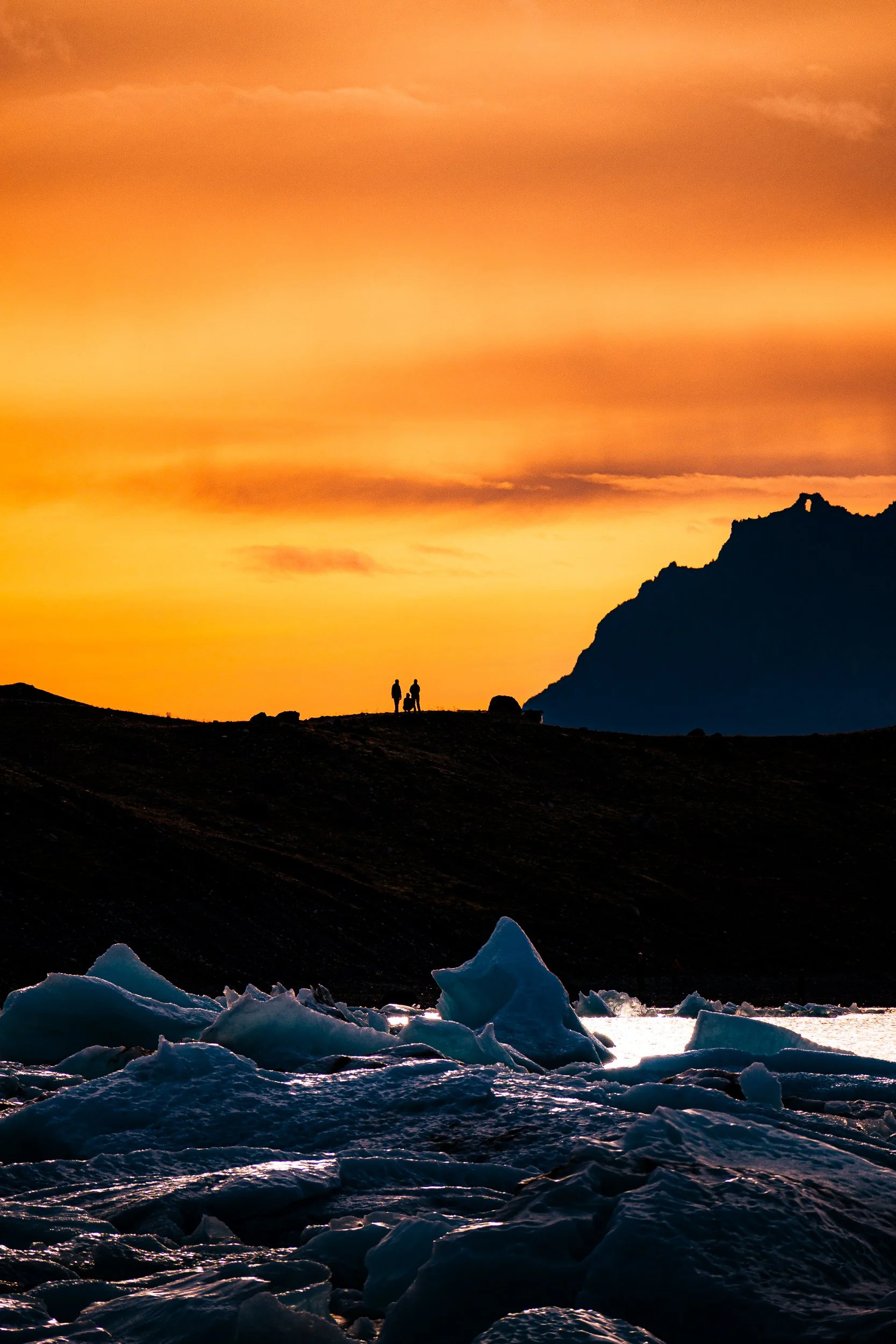 Two people silhouetted against an orange sunset beside an icy glacial lagoon, with a dark mountain ridge and floating blue ice chunks.