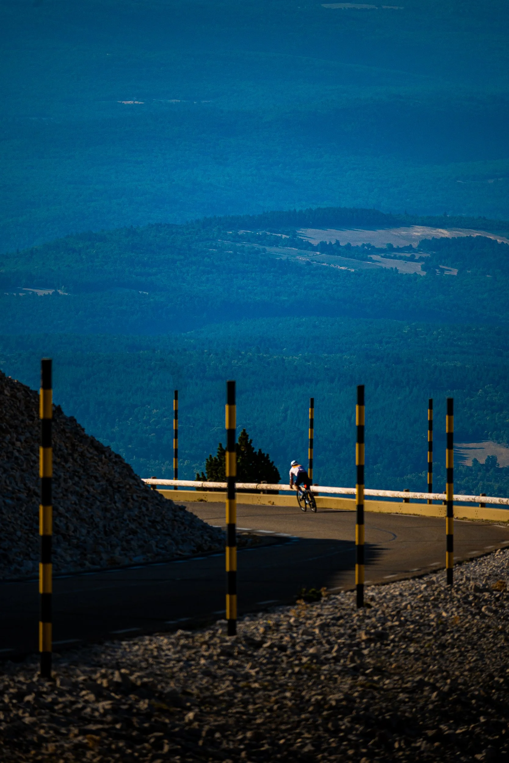 Cyclist riding along a mountain road lined with black-and-yellow posts, overlooking forested hills and farmland