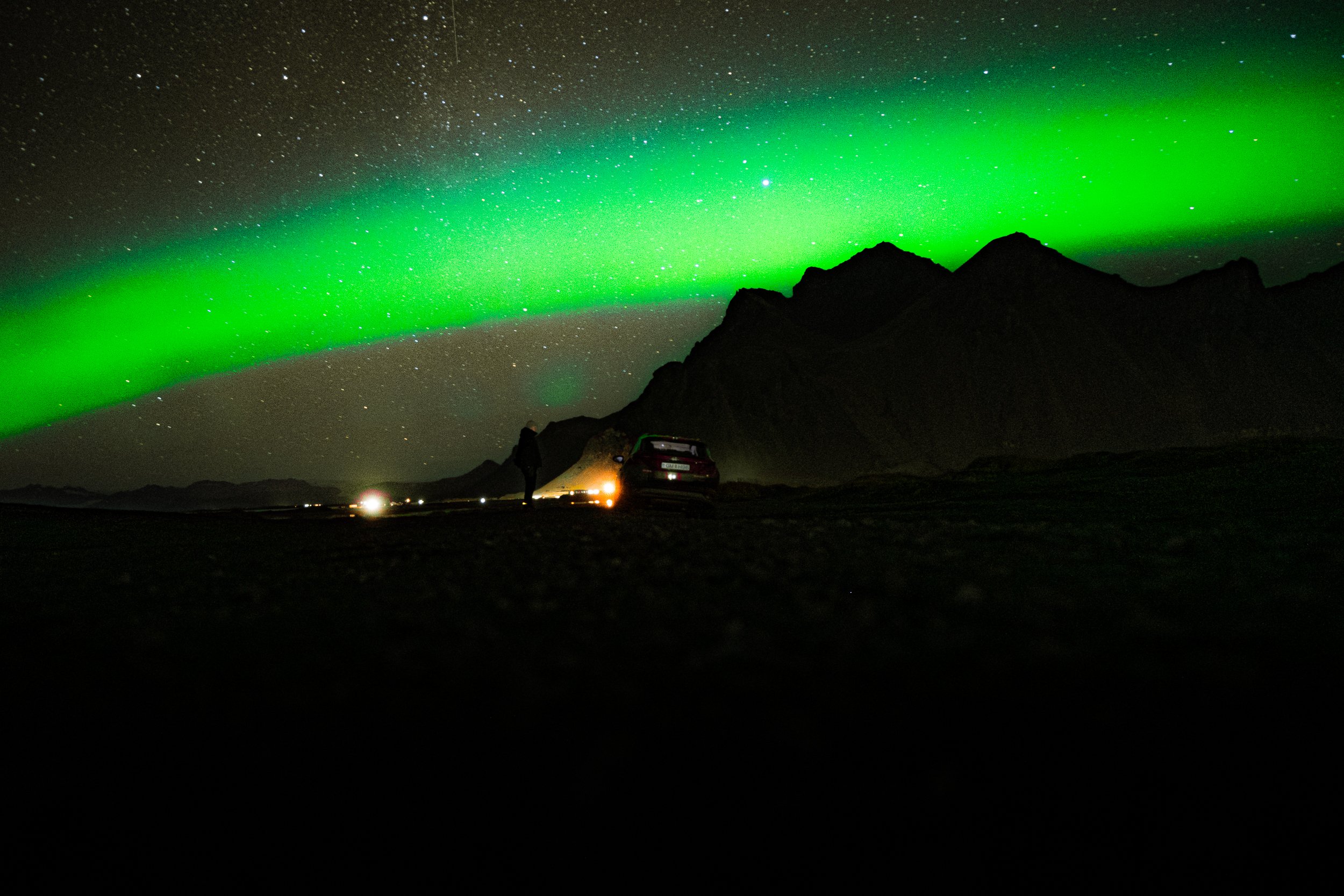 Bright green aurora borealis above a starry night sky and silhouetted mountains, with cars parked on a dark road.