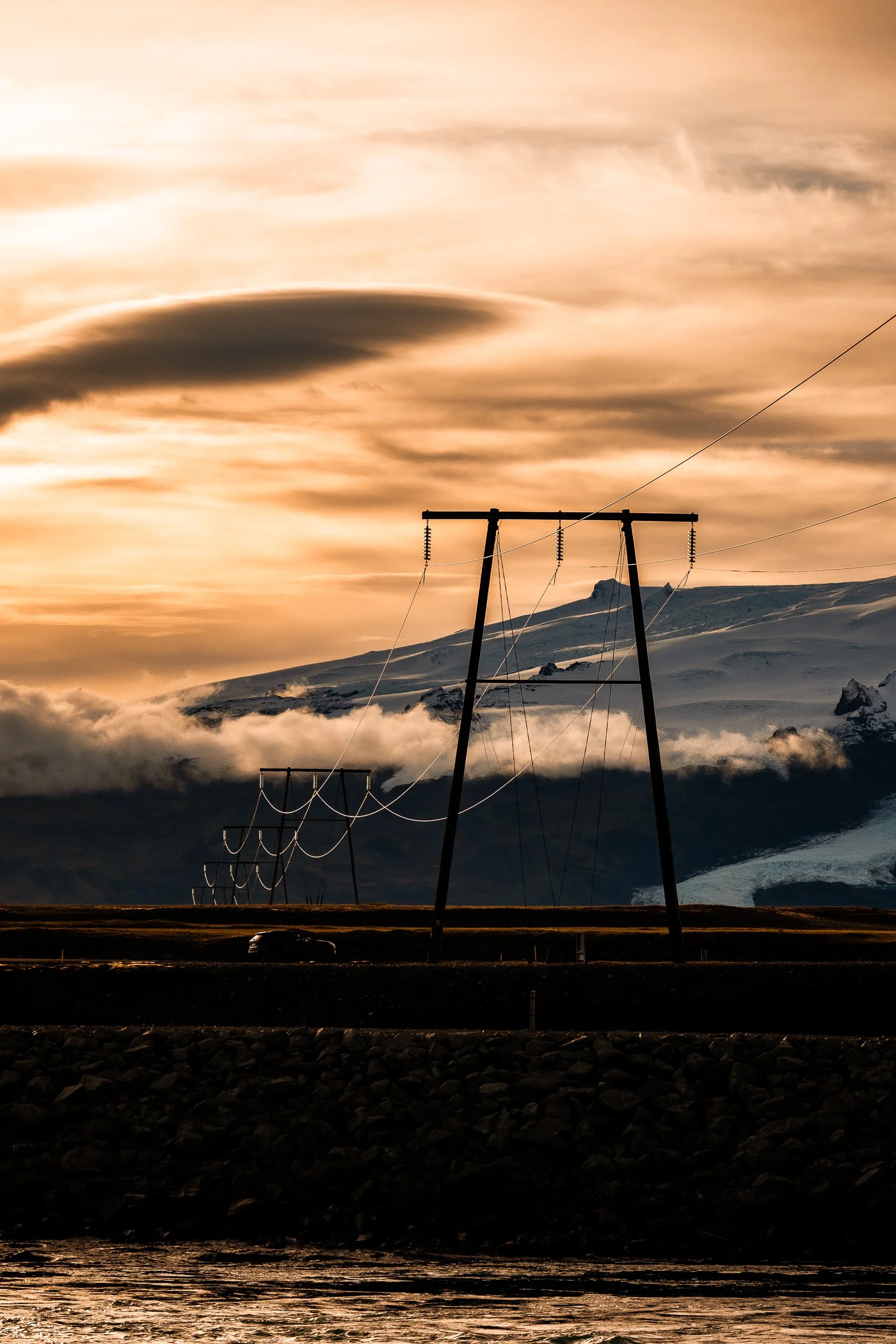 Power transmission towers and lines silhouetted against a glowing sunset over a snowy mountain and glacier