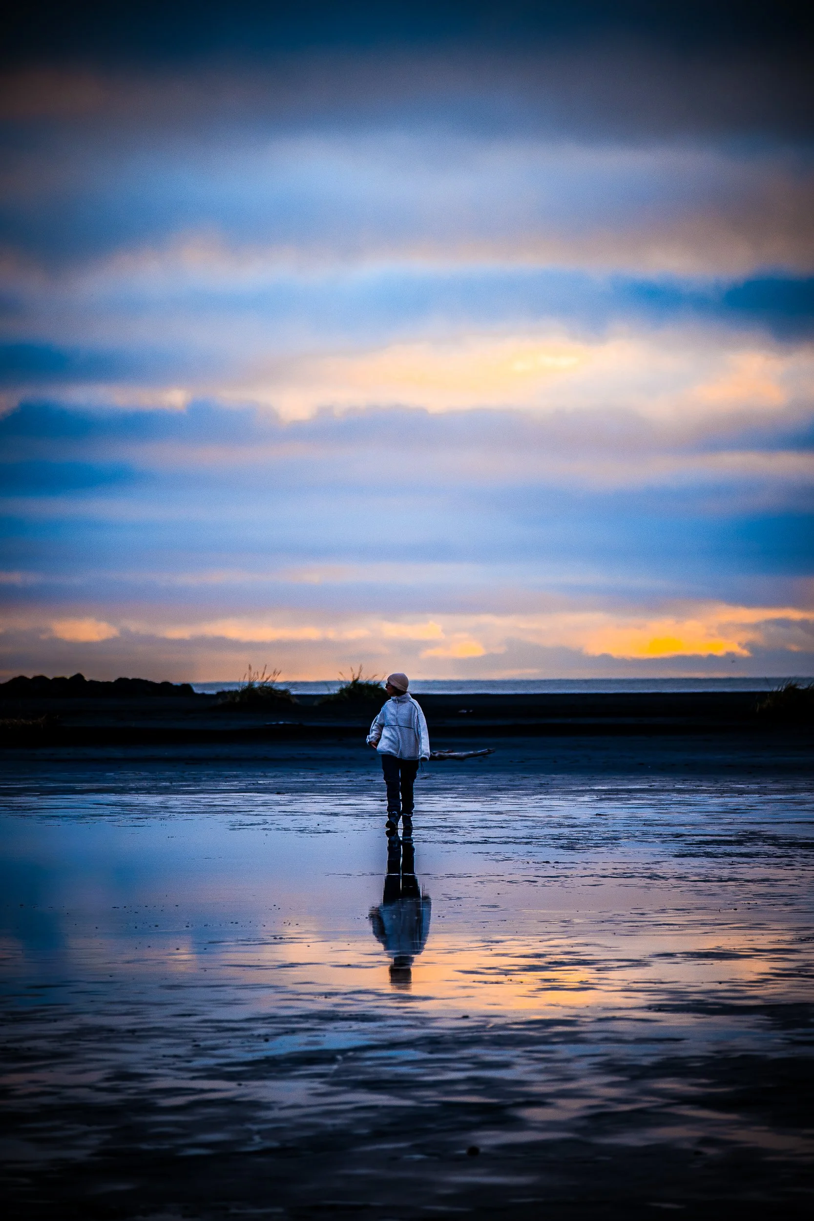 Person in a white jacket and hat standing in shallow ocean water at sunset, reflected beneath a colorful cloudy sky