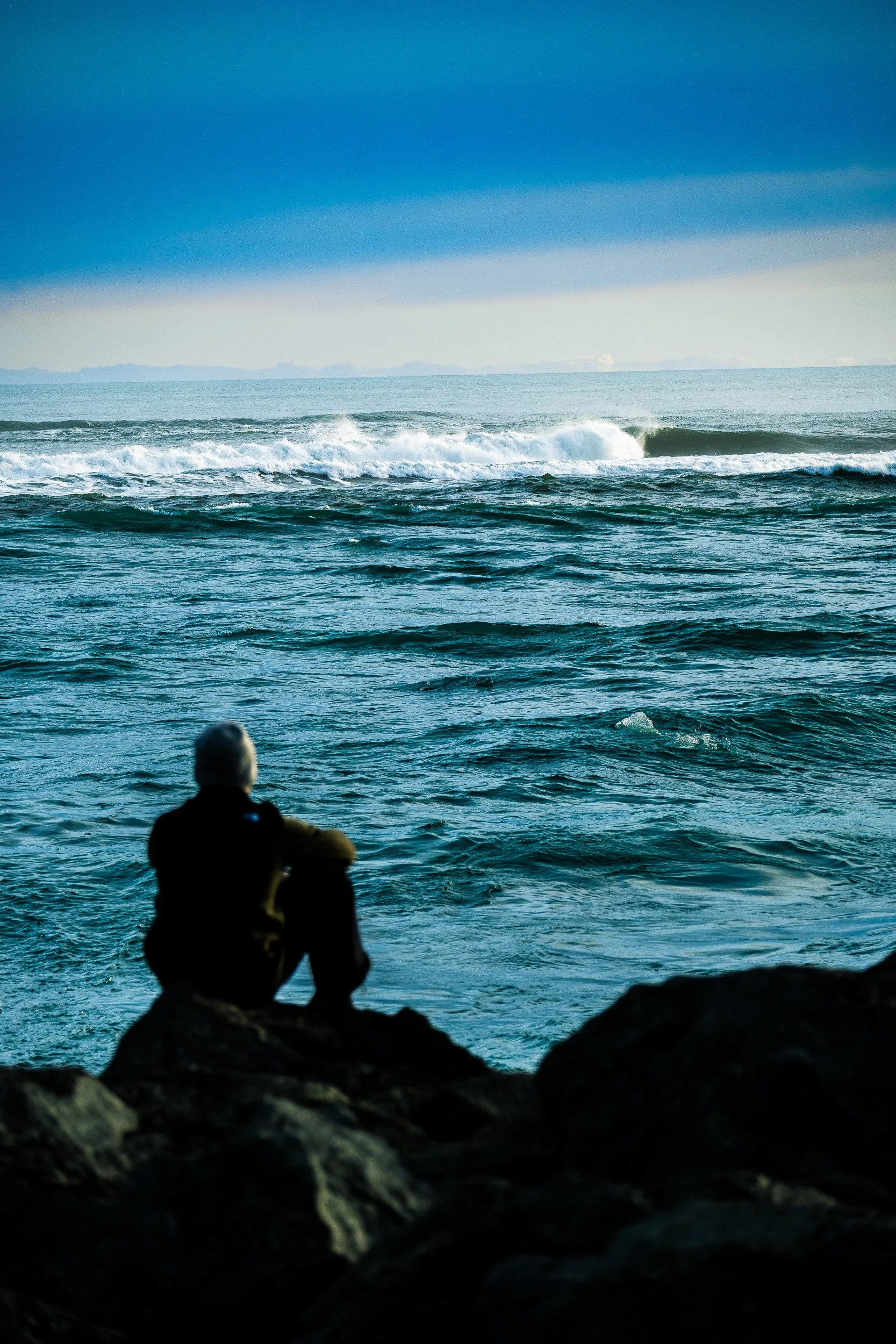 Silhouetted person sitting on coastal rocks and watching waves break across the blue ocean under a cloudy sky