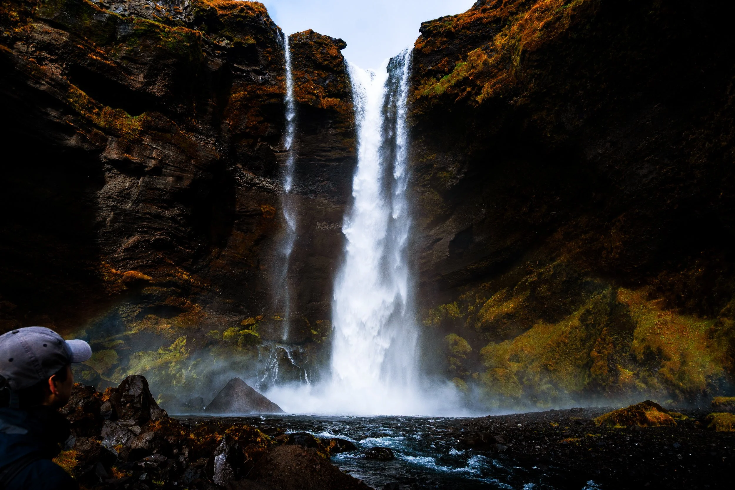 Person viewing a tall waterfall cascading through moss-covered volcanic cliffs into a rocky stream