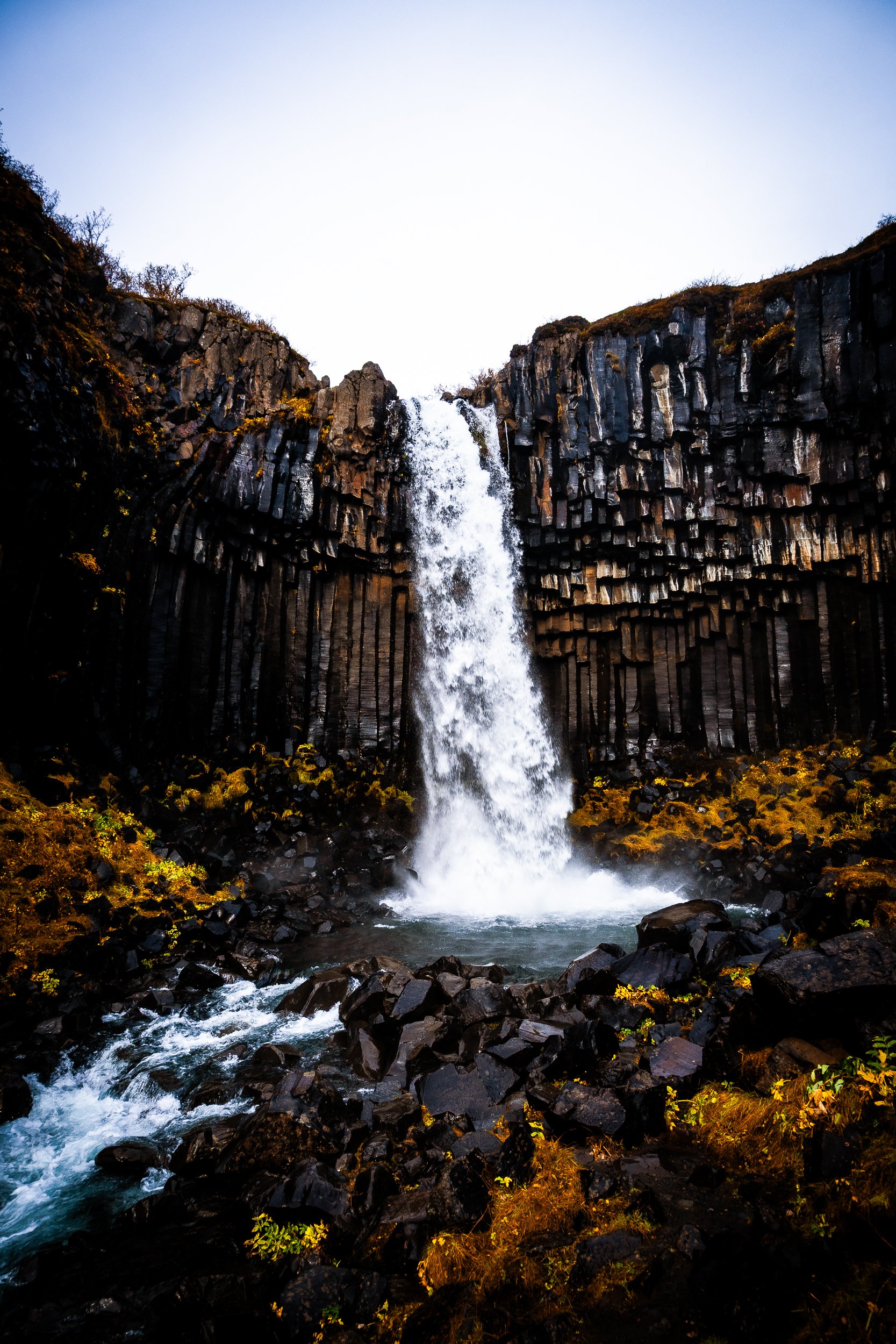 Waterfall cascading between dark basalt columns into a rocky pool surrounded by moss and autumn vegetation