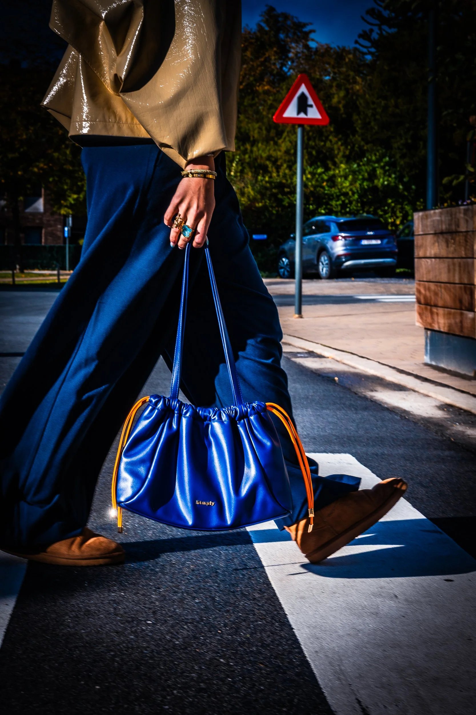 Person in beige trench coat and dark blue pants carrying a bright blue drawstring handbag across a city crosswalk