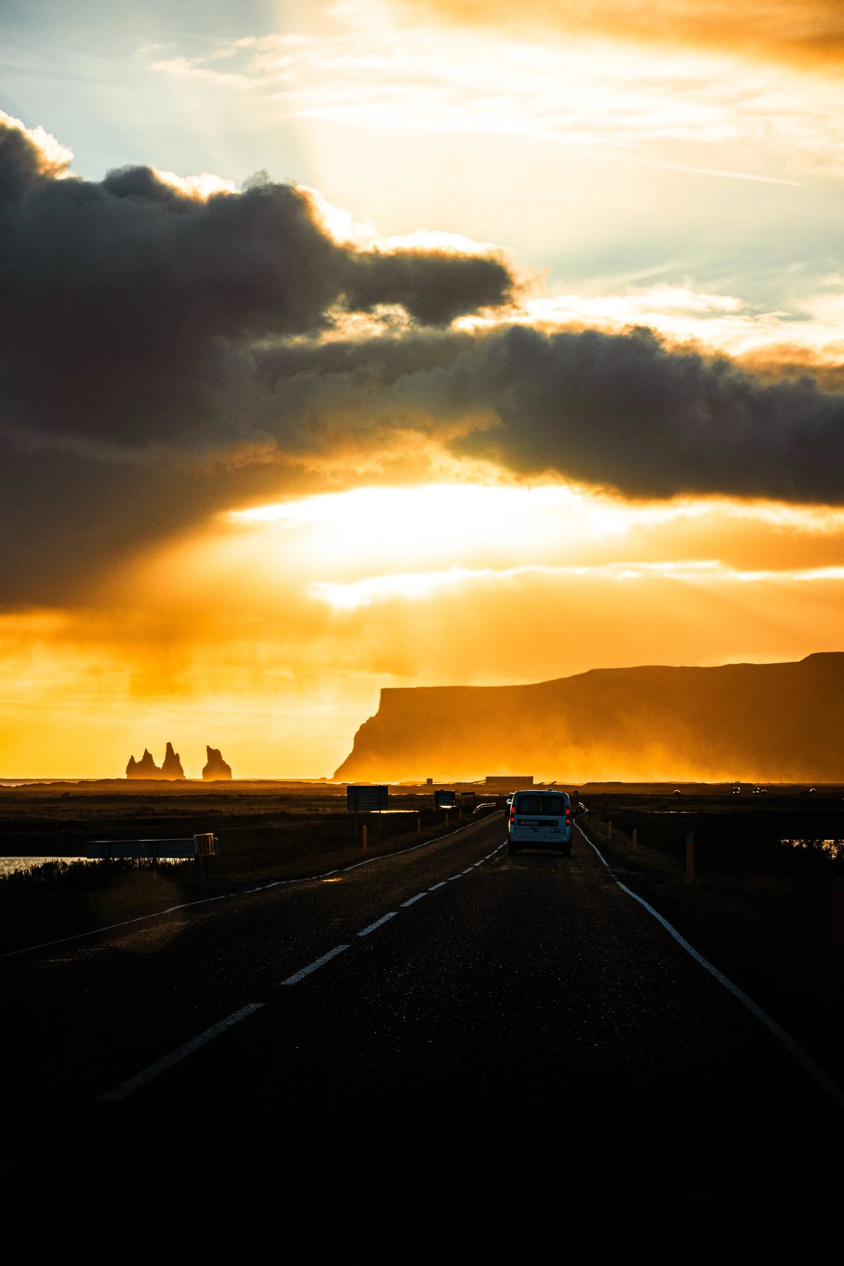 Sunset over Iceland’s volcanic coastline, with dramatic clouds, sea stacks, cliffs, and a van driving along a coastal road.