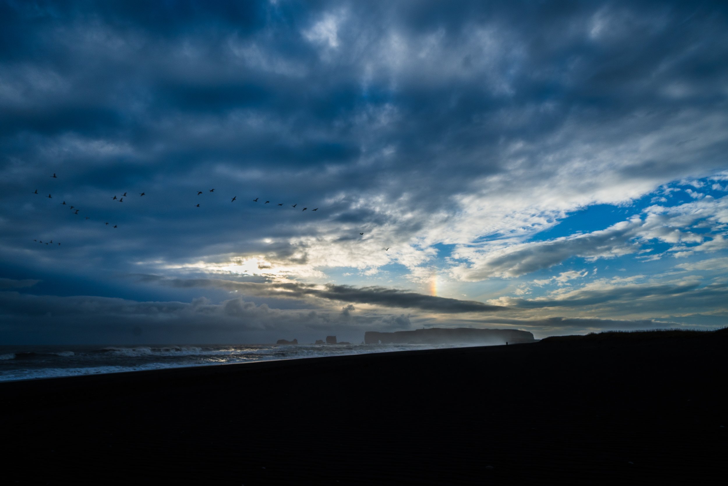 Black sand beach with crashing waves, rock formations, seabirds, and a rainbow beneath dramatic storm clouds