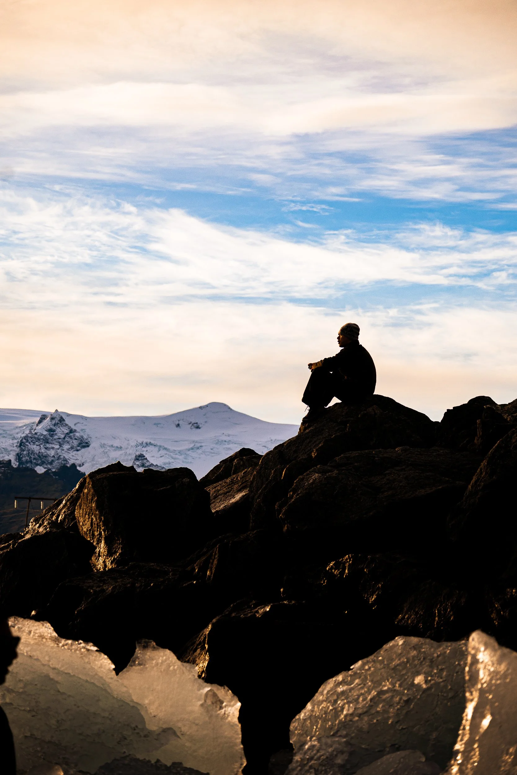Silhouetted hiker sitting on dark rocks overlooking a snow-covered mountain beneath a pastel sunset sky