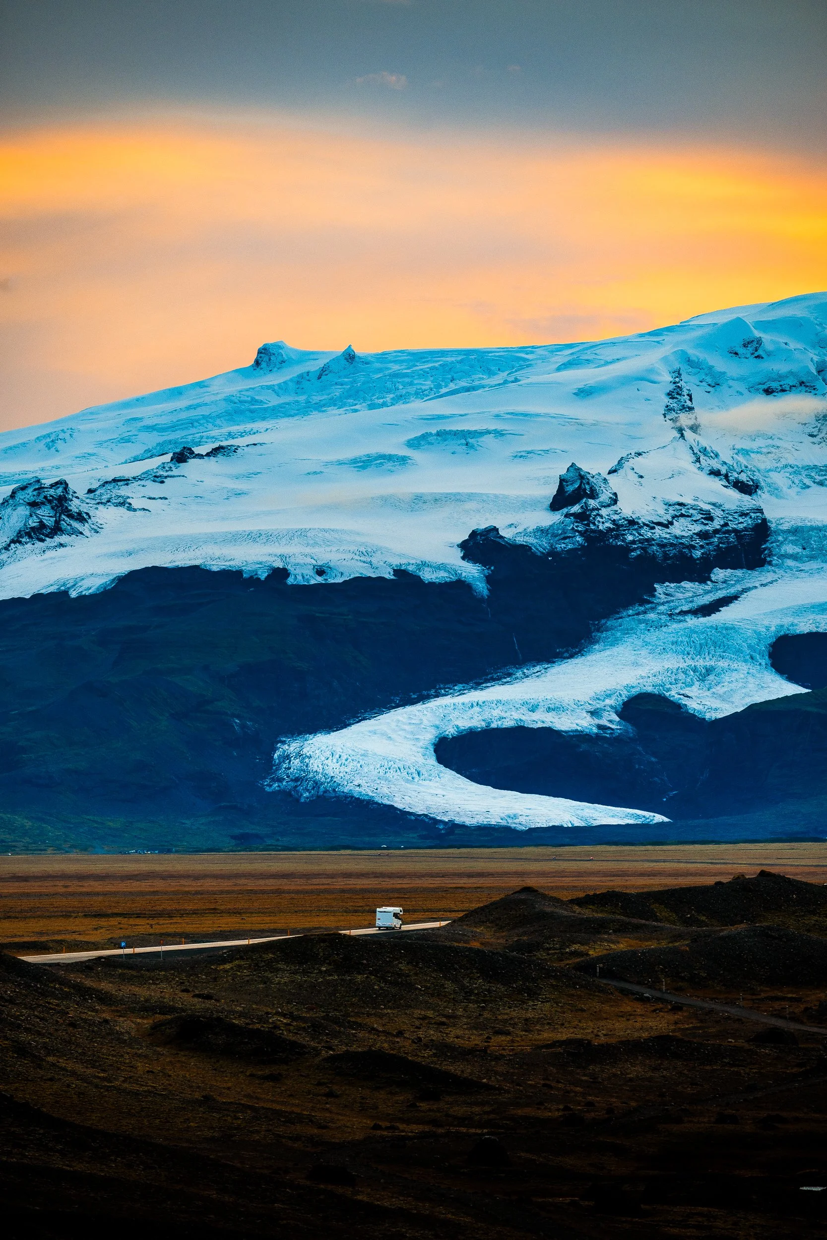 Snow-covered glacier and volcanic mountain above a road with a vehicle at sunset