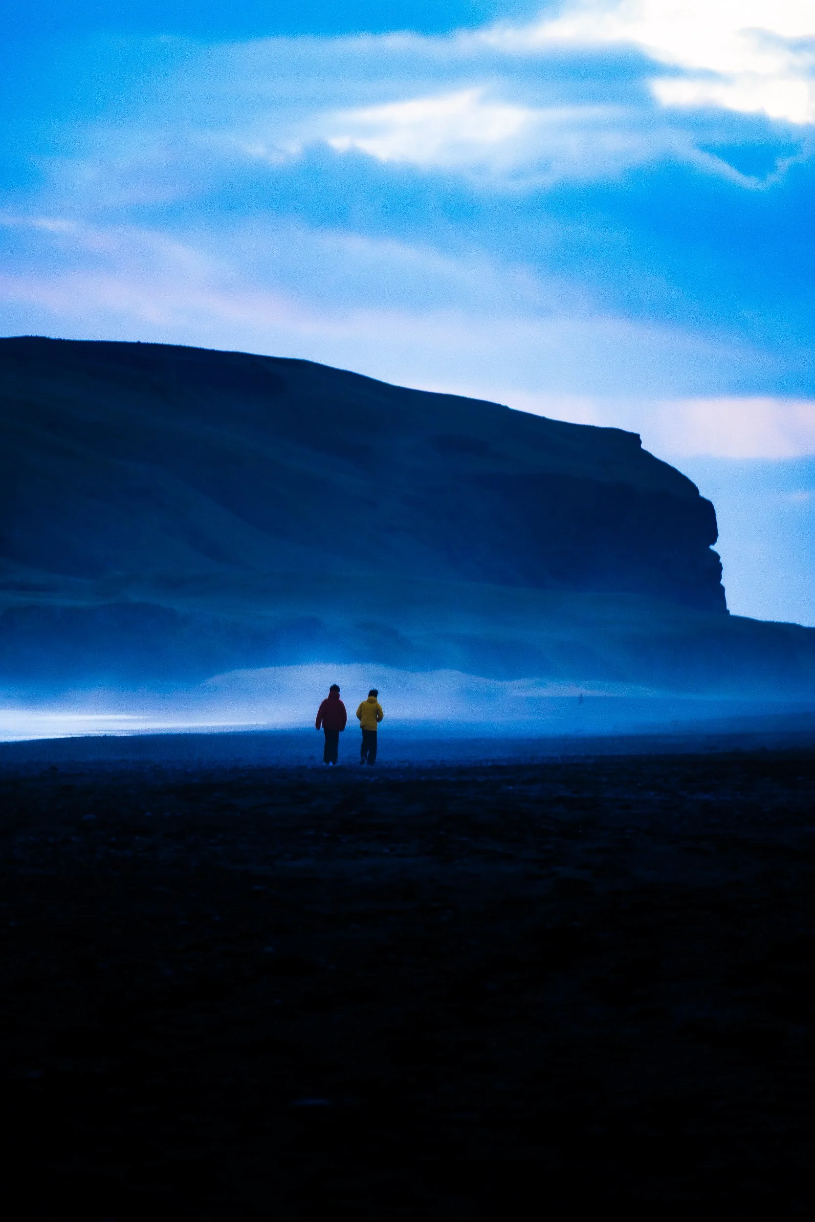 Two hikers walk beneath a dramatic blue sky toward a rugged mountain cliff.