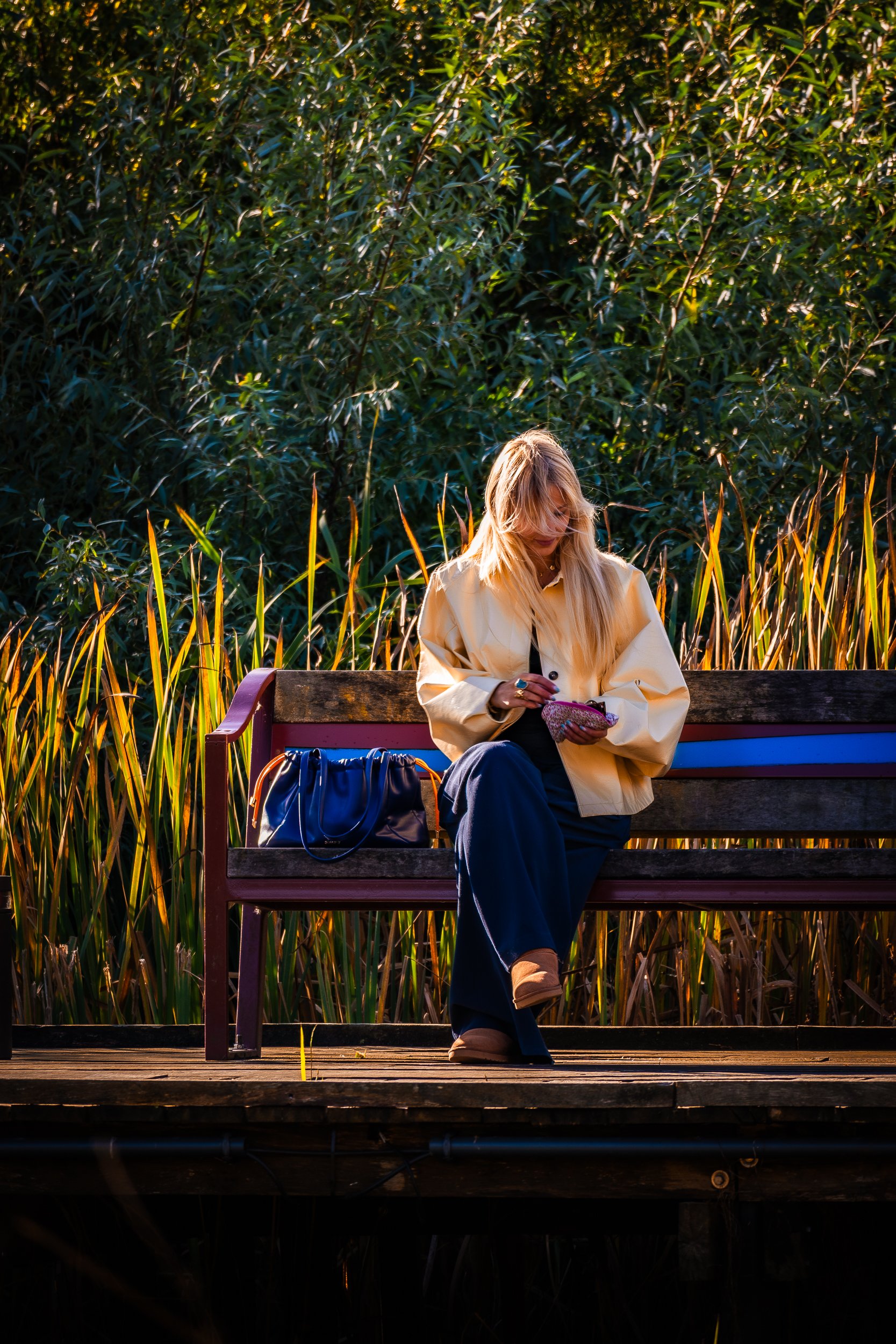 Blonde woman in a cream coat sitting on a bench beside a pond, holding a pink clutch with a blue handbag nearby.