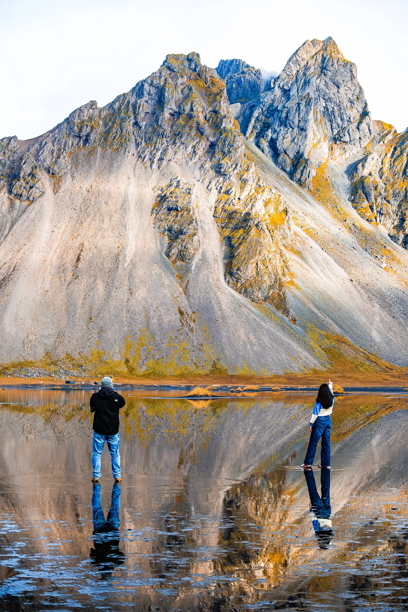 Two tourists photographing rugged golden mountain peaks reflected in a calm alpine lake
