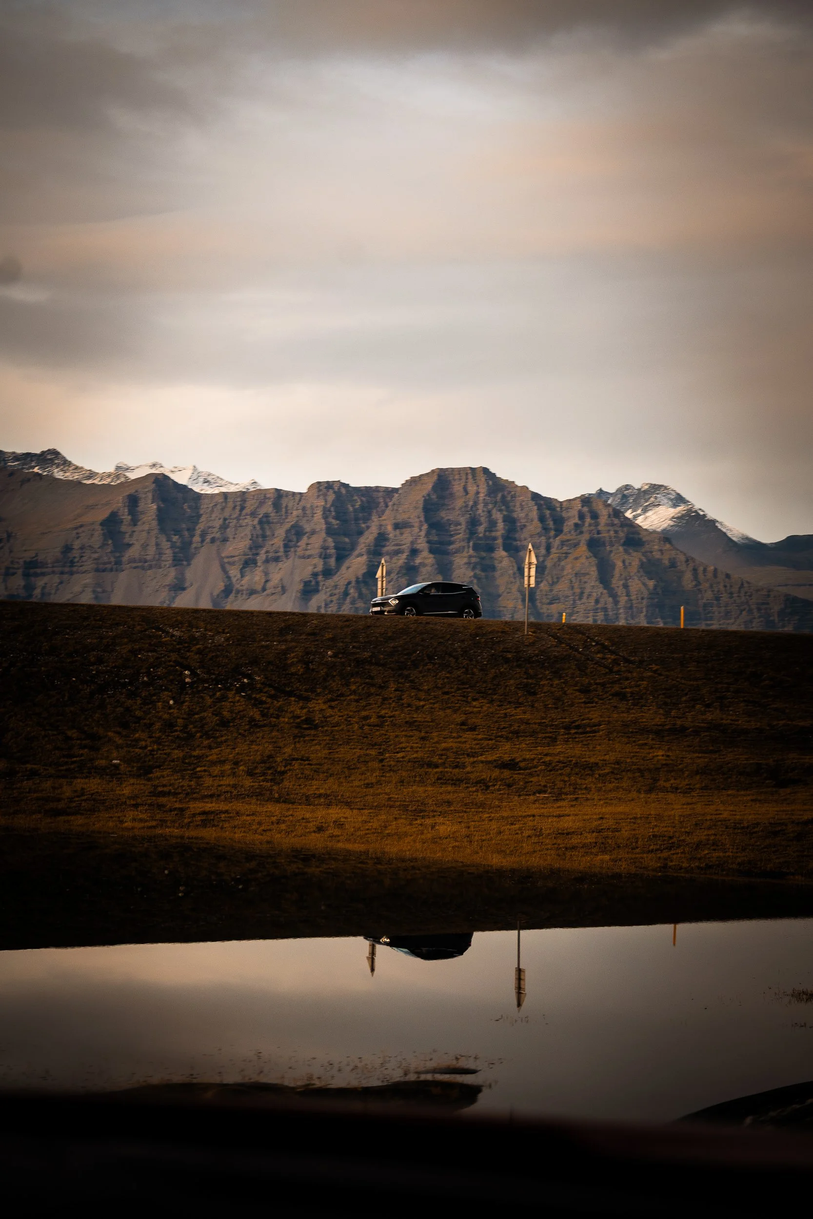 Black car driving past rugged snow-capped mountains beside a reflective lake at sunset