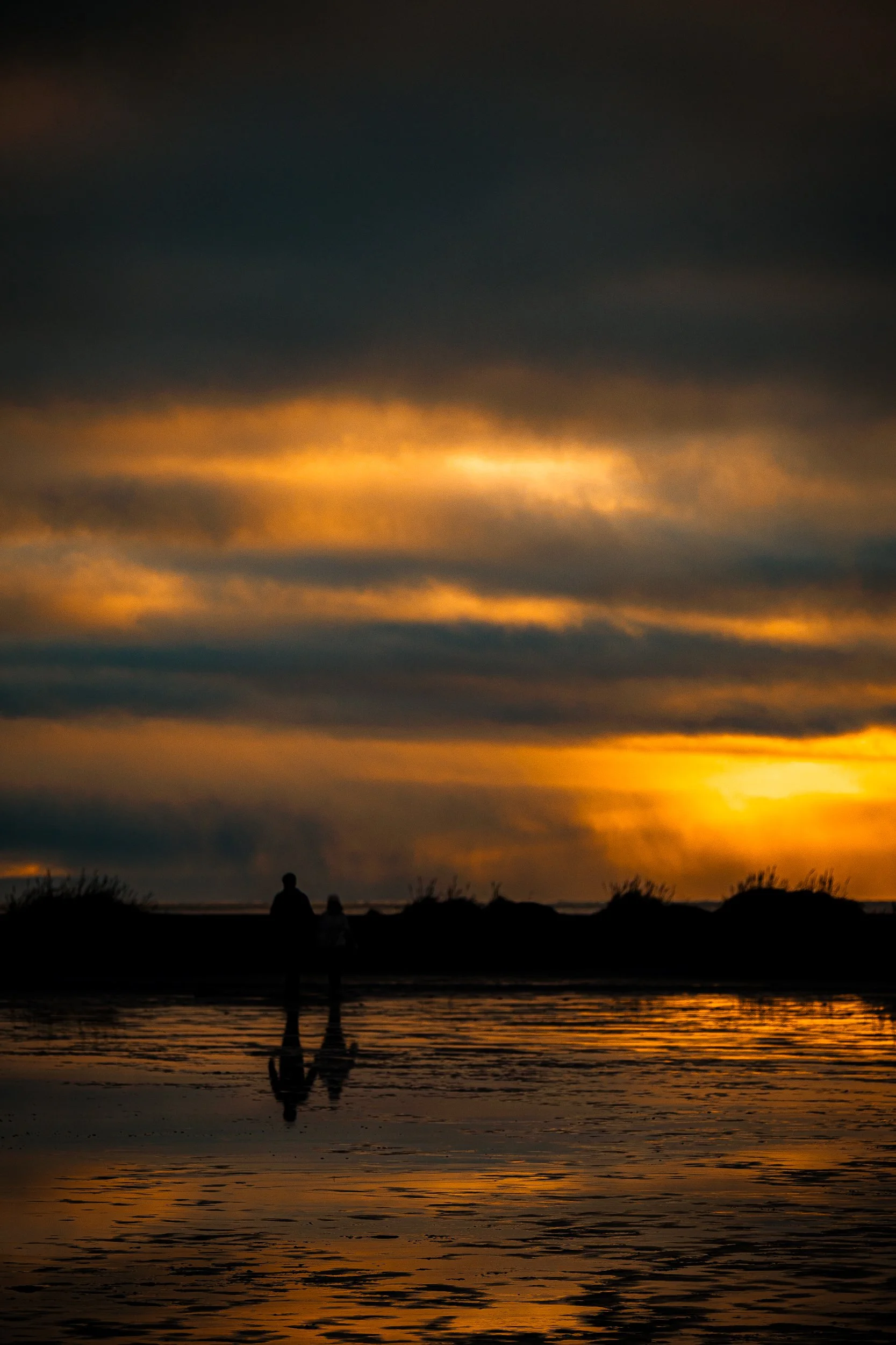 Silhouetted people standing in shallow water beneath dramatic orange sunset clouds