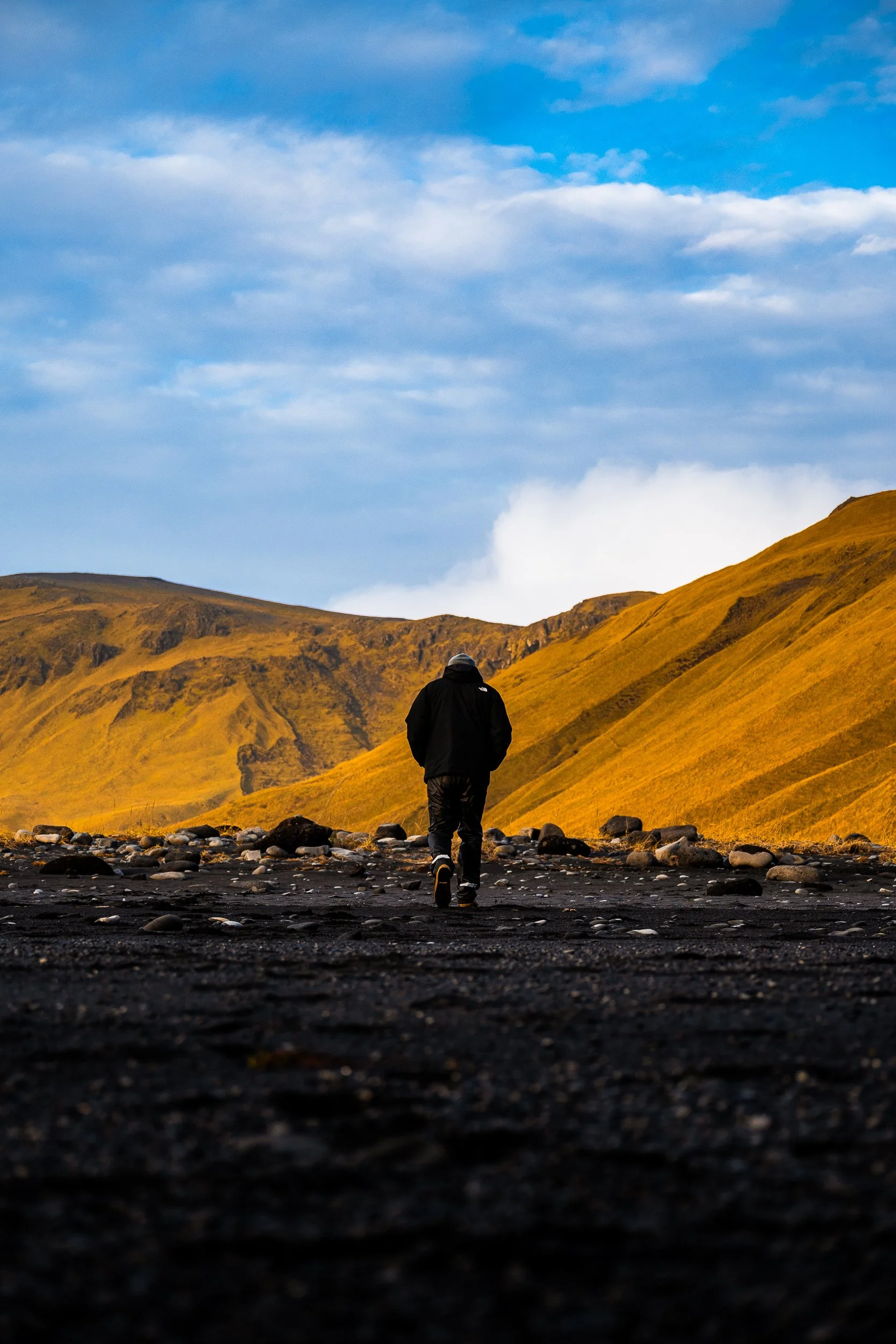 Hiker in black clothing walking across a rocky valley beneath golden mountains and a cloudy blue sky