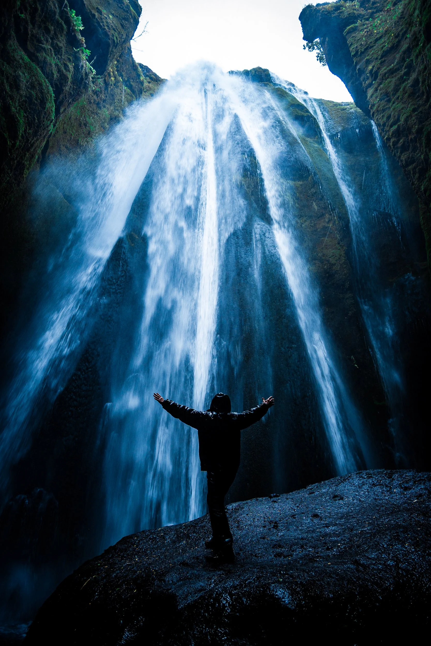 Silhouetted person with arms outstretched beneath a powerful waterfall in a moss-covered canyon