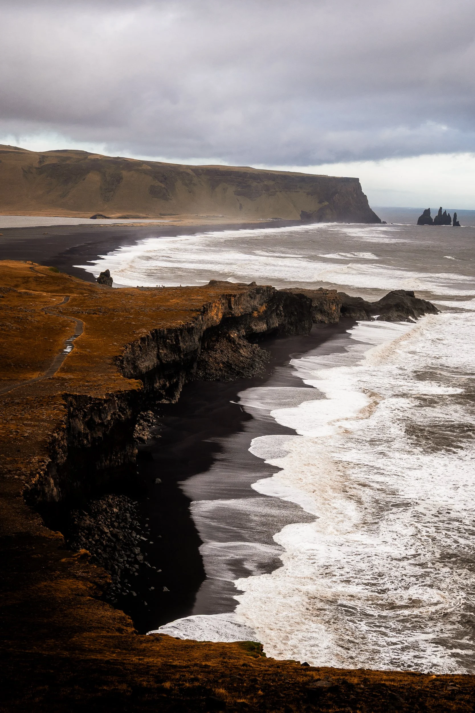 Black sand beach with basalt sea cliffs, crashing waves, and offshore rock formations beneath storm clouds