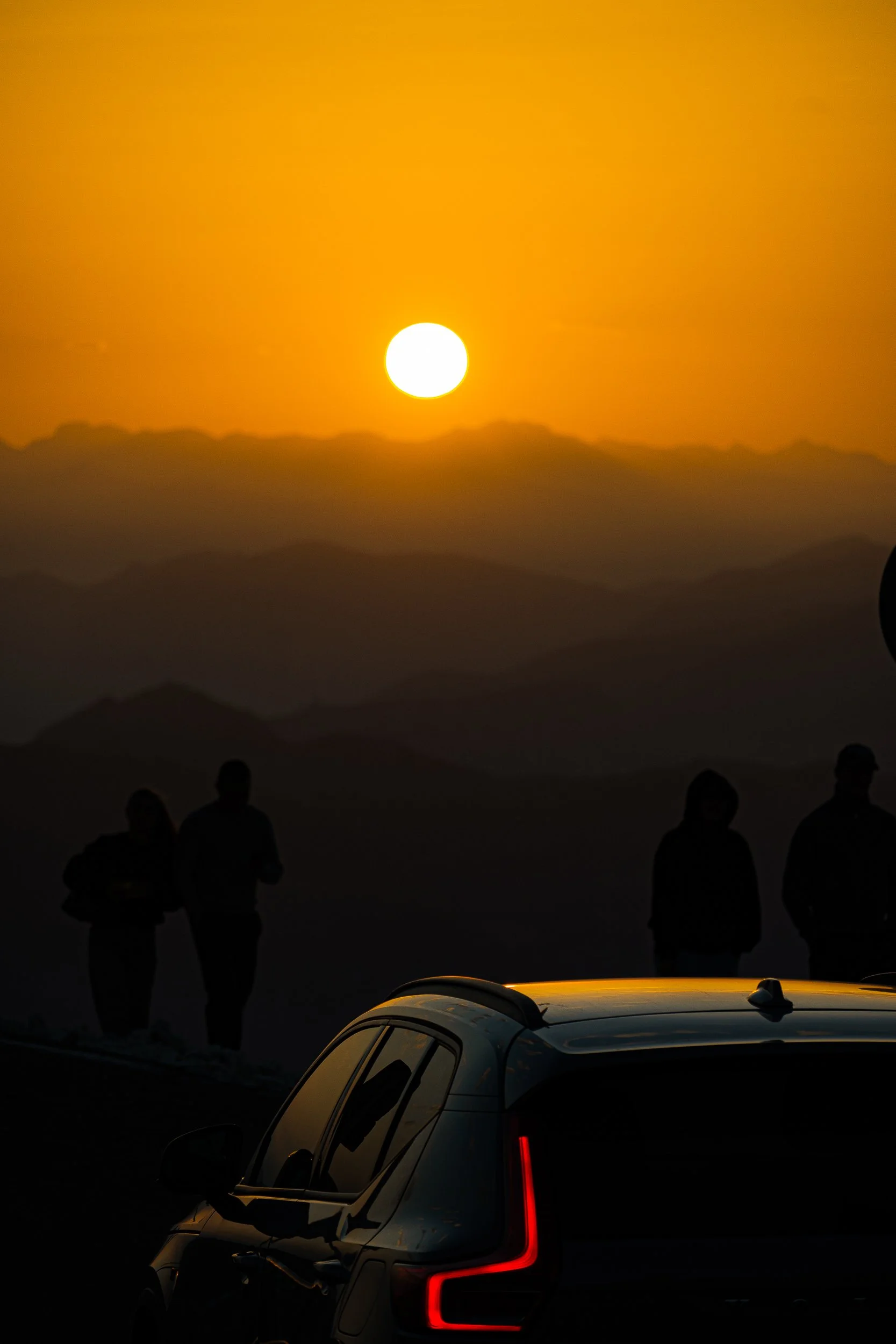 Silhouetted people and an SUV against a bright orange sunset over the mountains
