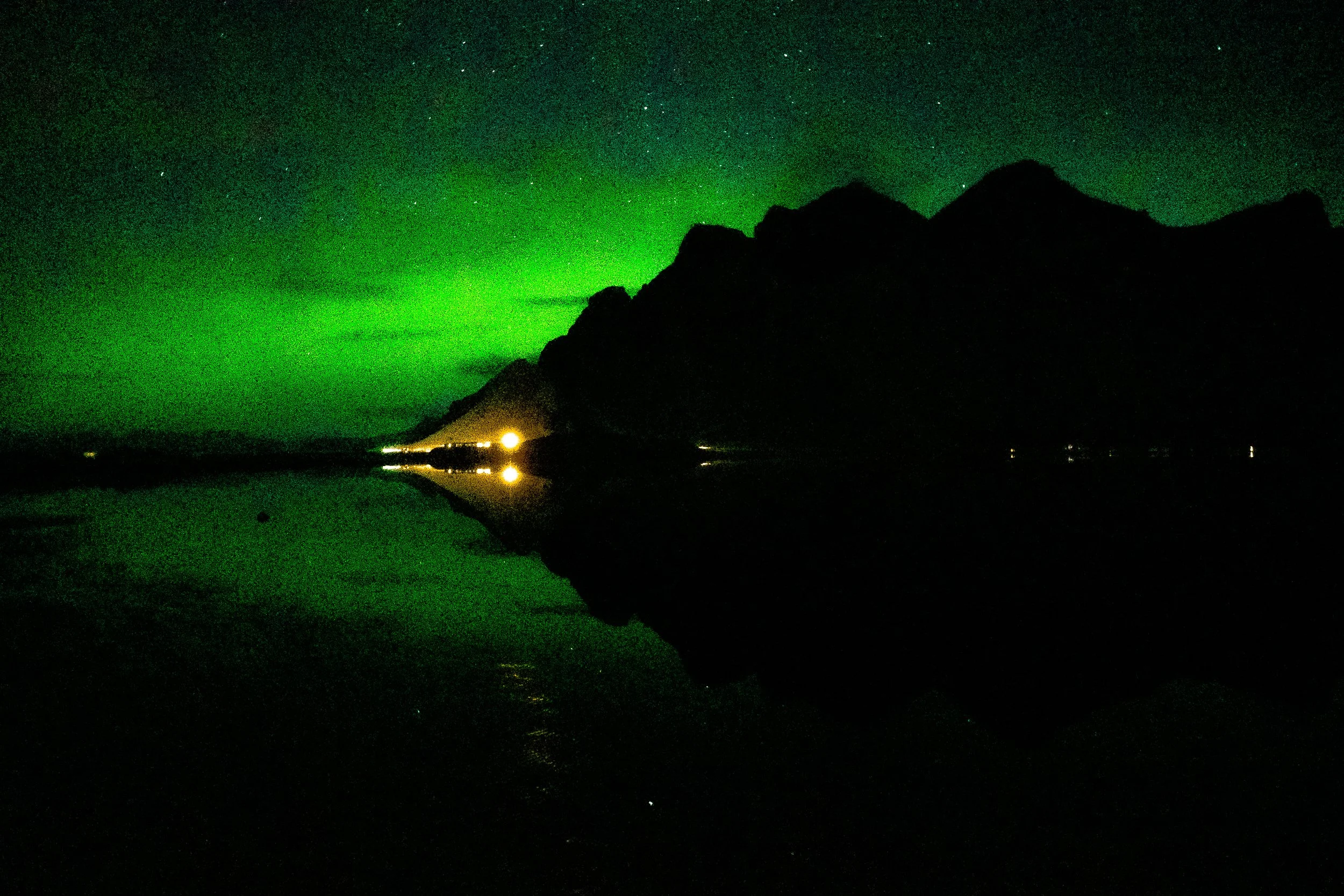 Nighttime mountain silhouette beneath a vivid green starry sky, with warm lights reflecting on a calm lake