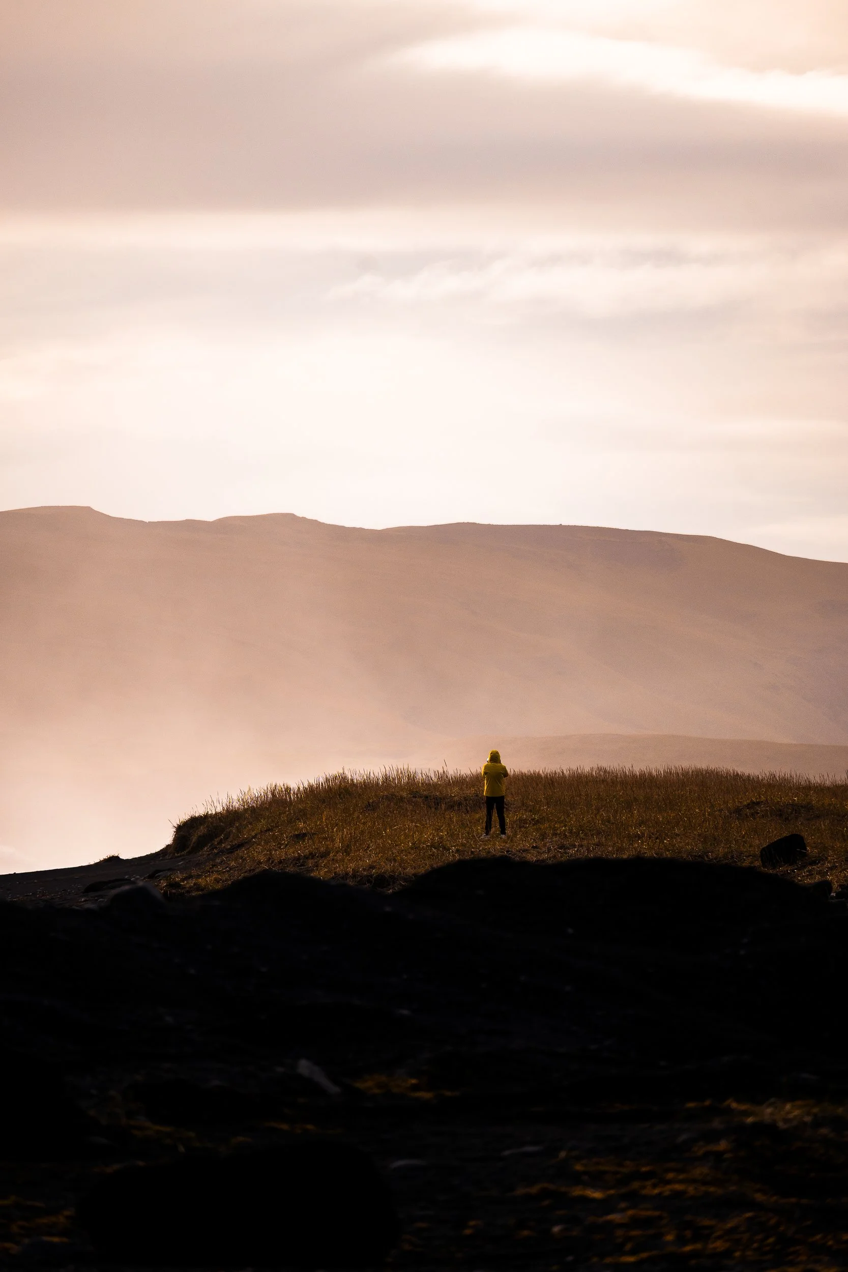 Person in a yellow jacket standing on a grassy ridge overlooking expansive sand dunes beneath a cloudy sky