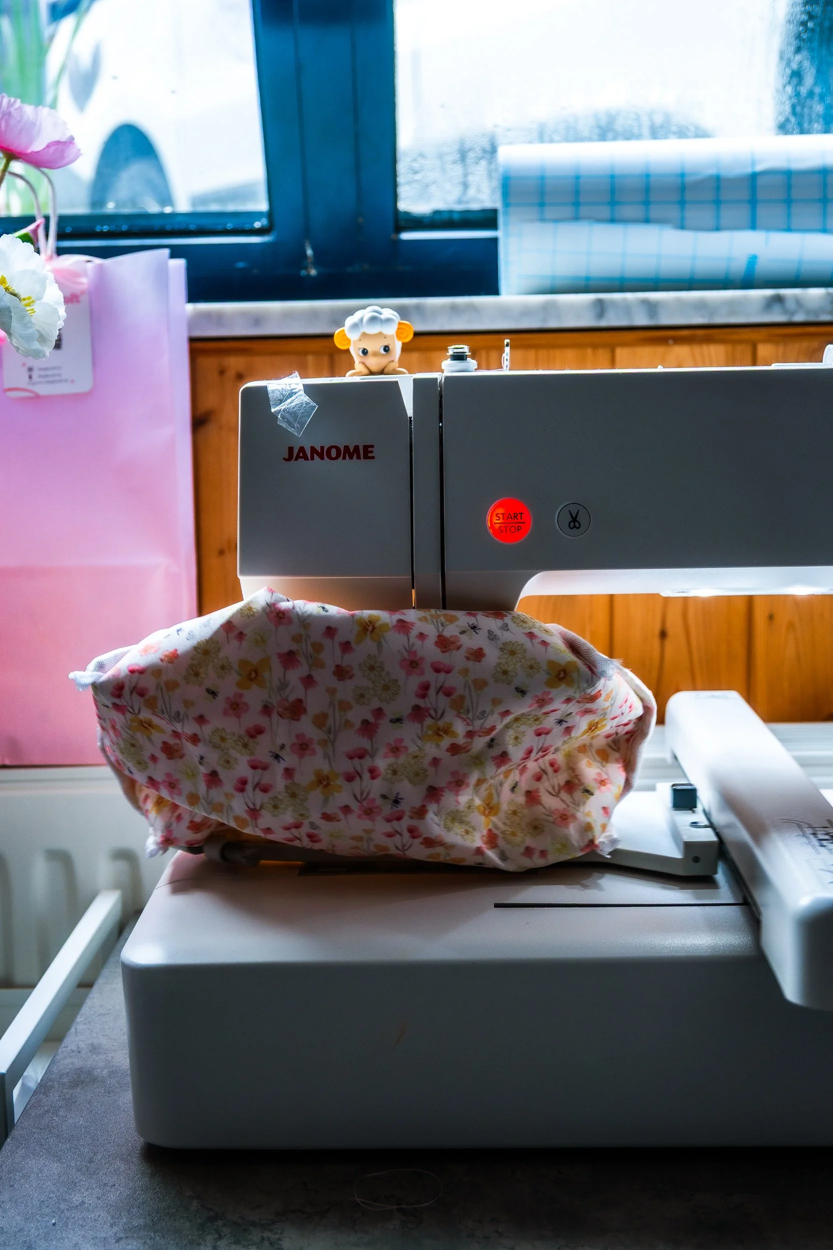 Janome sewing machine with floral fabric, pink flower vase, and small doll by a window
