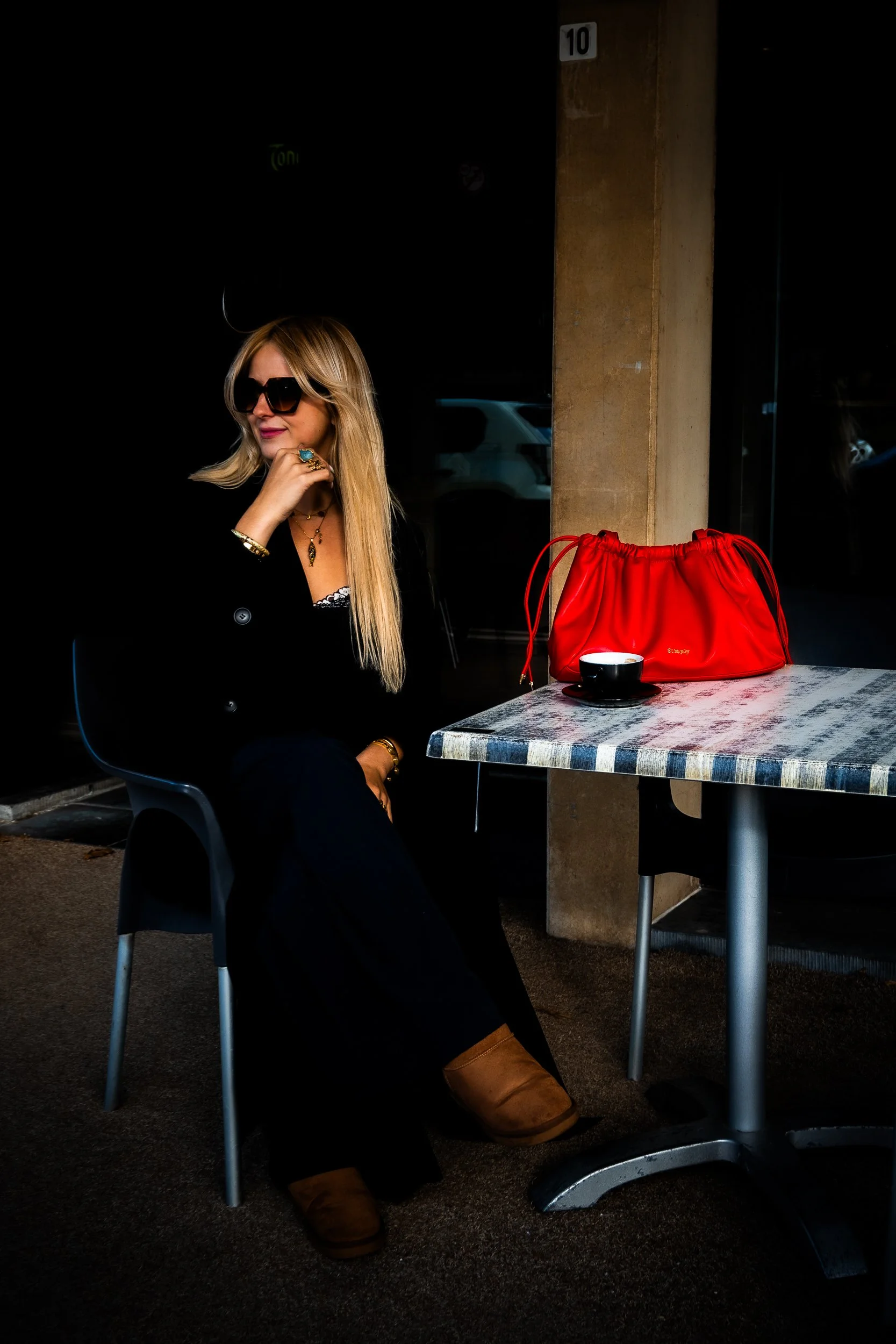Blonde woman wearing sunglasses sits at an outdoor café table with a red handbag and cup of coffee.