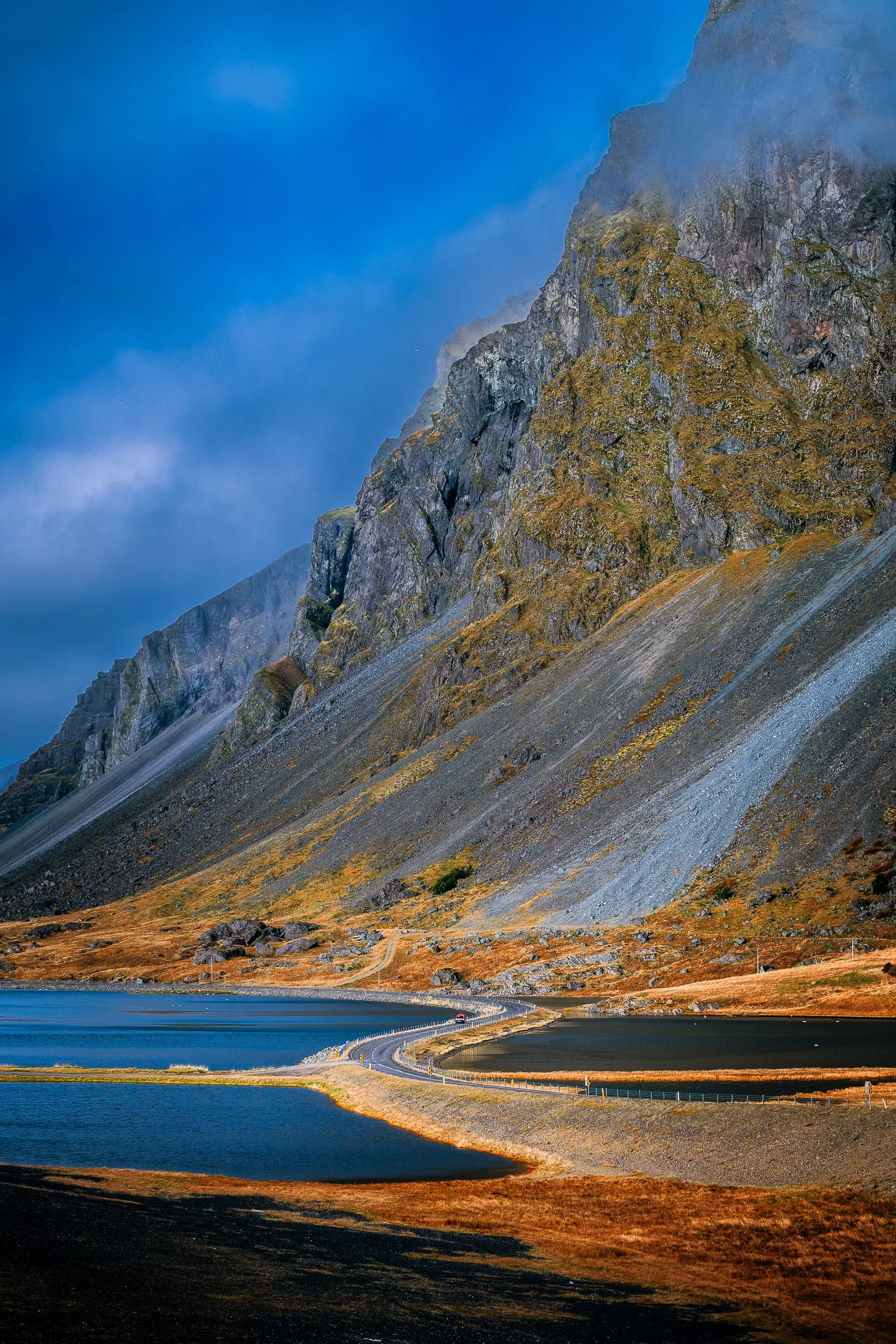 Scenic mountain road winding between blue alpine lakes beneath steep, rugged, moss-covered cliffs.