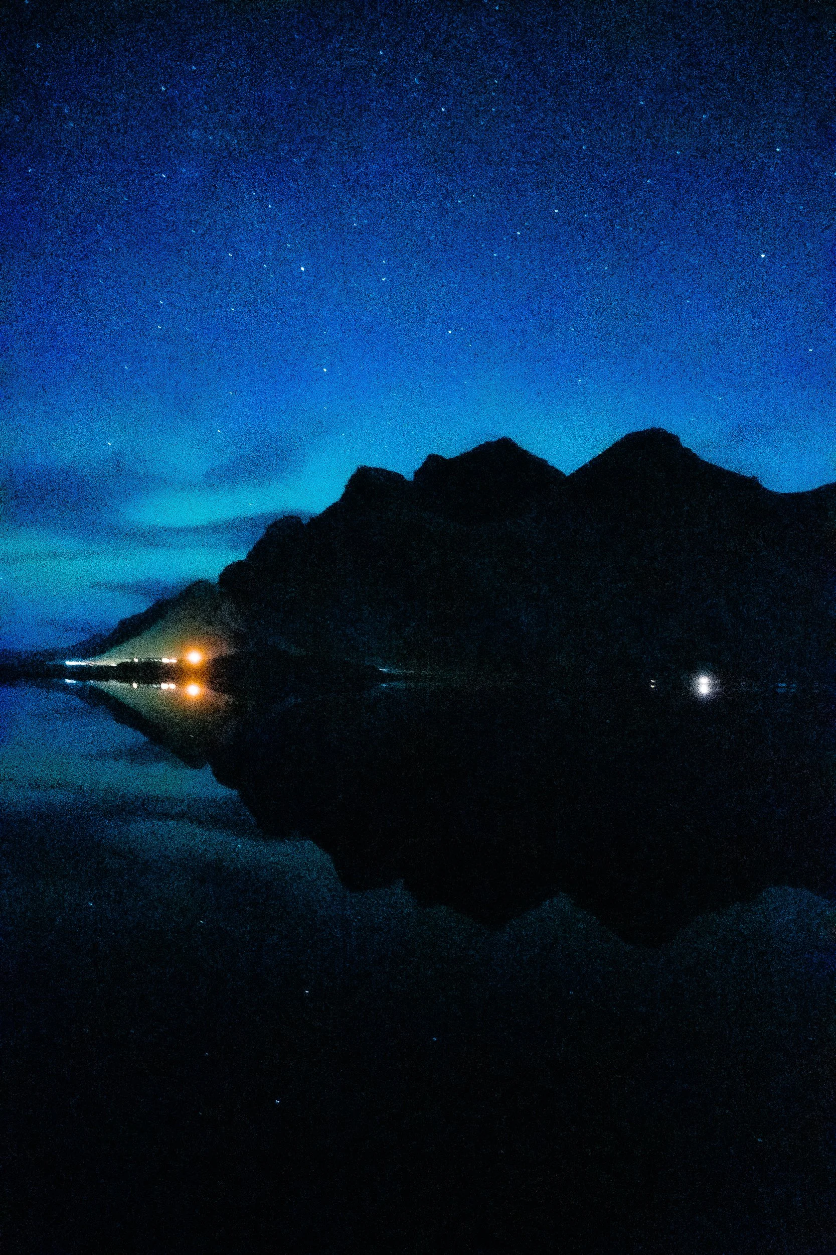 Starry night sky over a jagged mountain silhouette and dark reflective water with scattered shoreline lights