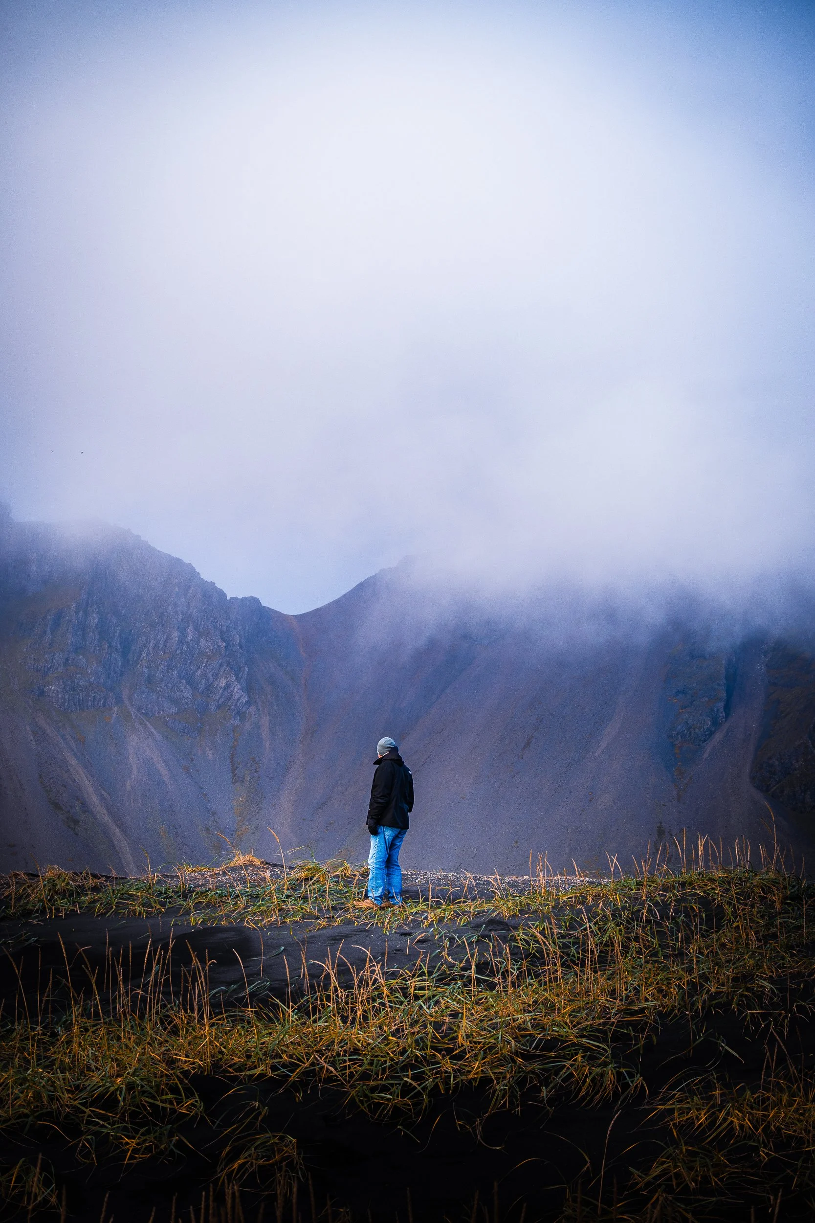 Hiker in a black jacket standing in a misty mountain valley surrounded by rugged cliffs and tall grass