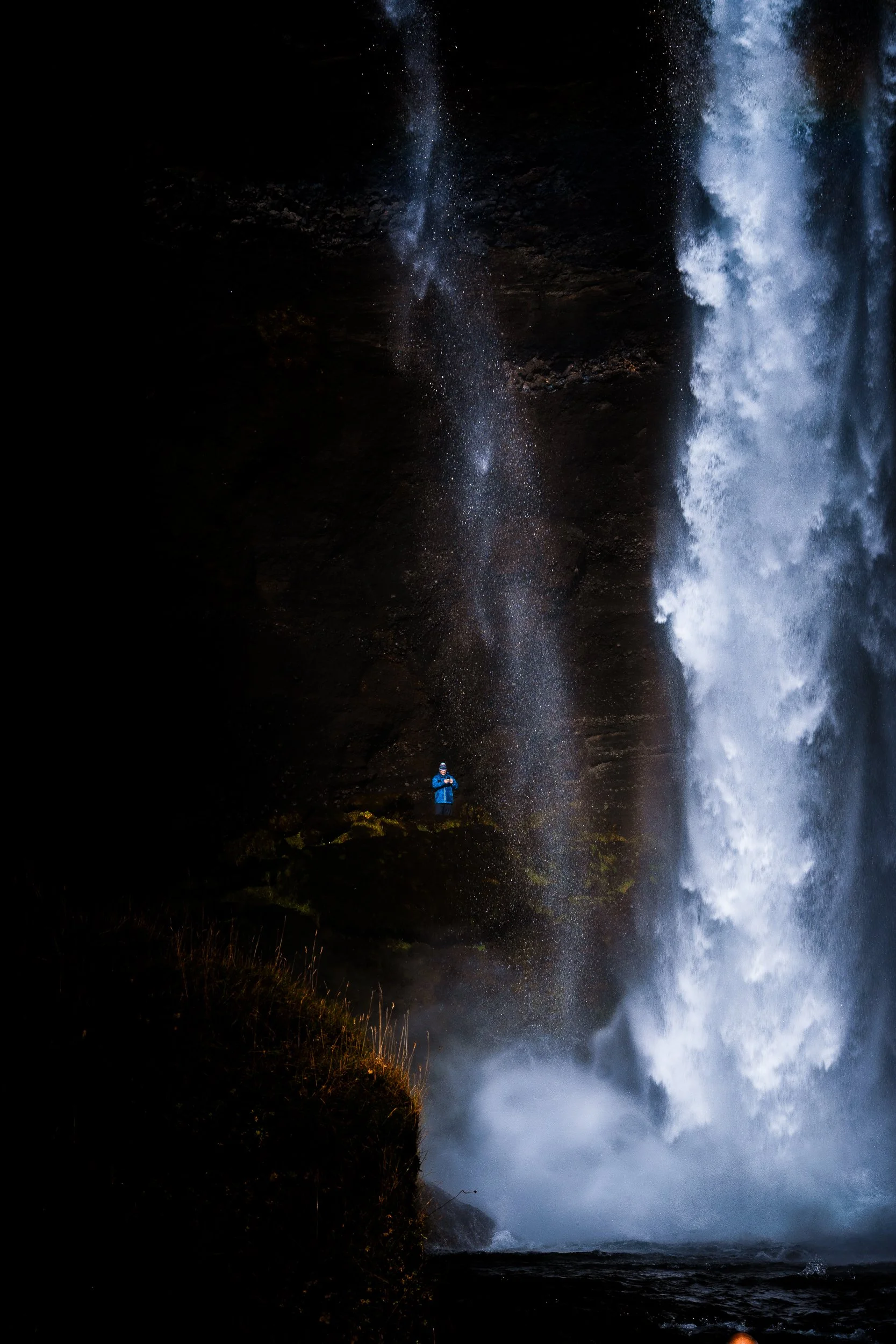 Hiker in a blue jacket standing beside a powerful waterfall in a dark rocky gorge