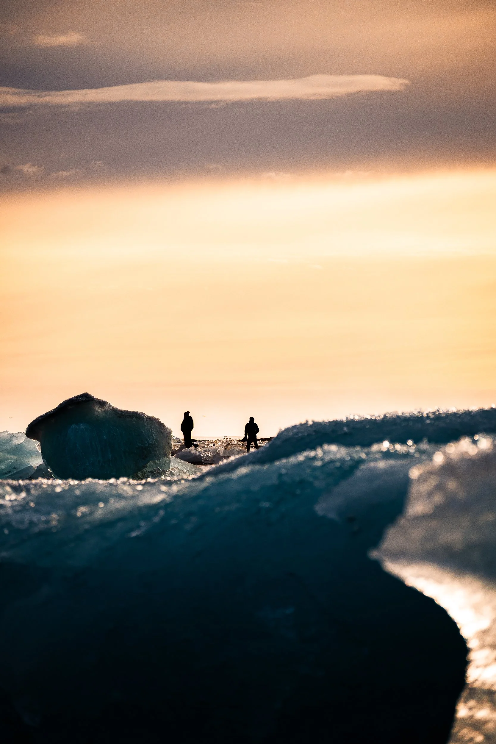 Two silhouetted people standing among blue icebergs on a calm ocean at sunset