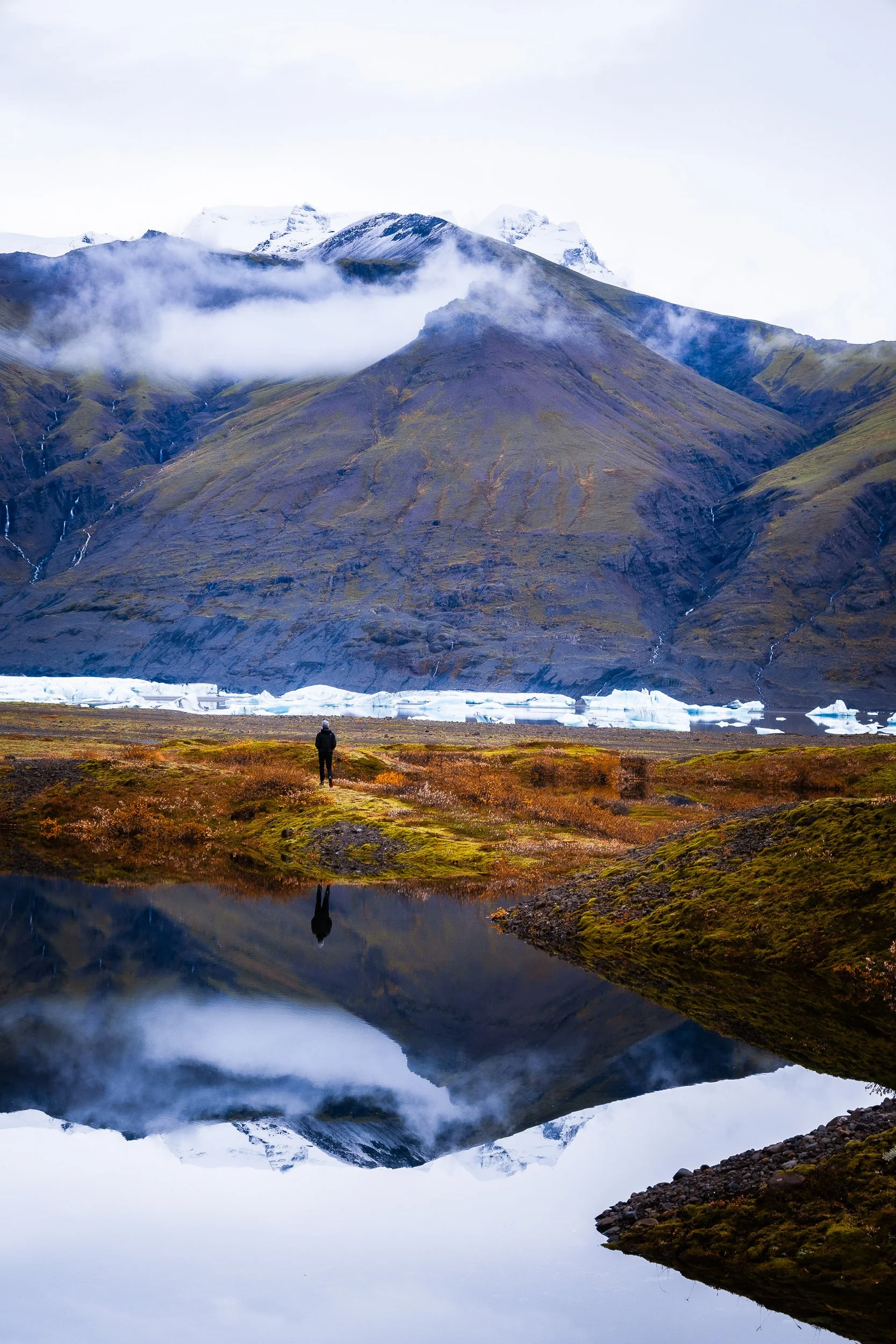 Traveler overlooking a glacial lagoon with floating icebergs, misty snow-capped mountains, and reflections in Iceland