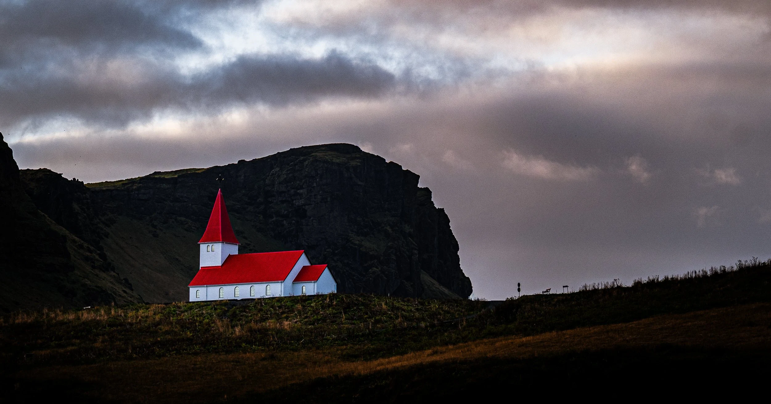White church with bright red roofs beneath a dramatic cloudy sky and dark Icelandic cliffs