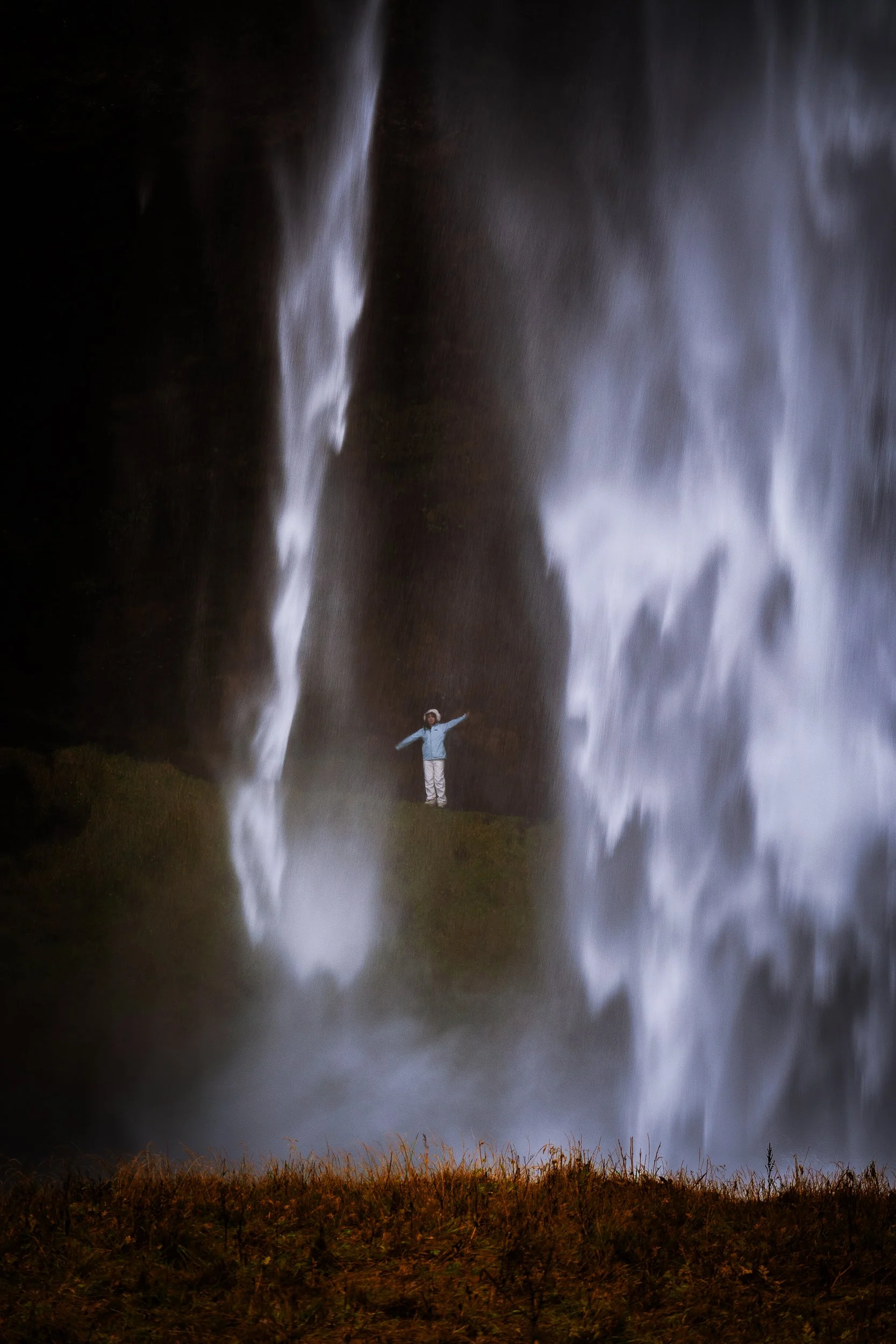 Person standing with arms outstretched between two tall waterfalls in a grassy landscape