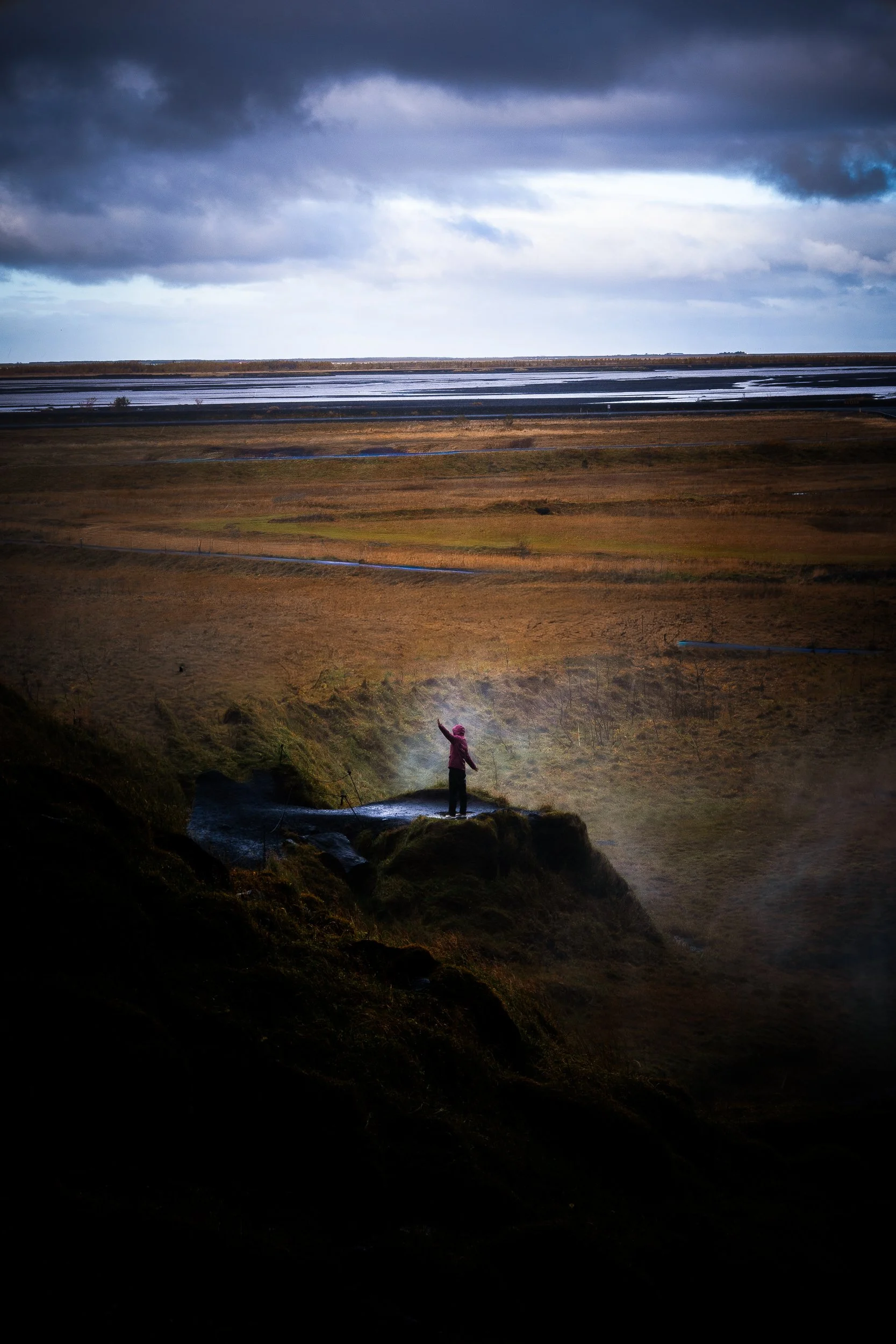 Person in a pink jacket standing on a cliff above a coastal wetland under dramatic storm clouds