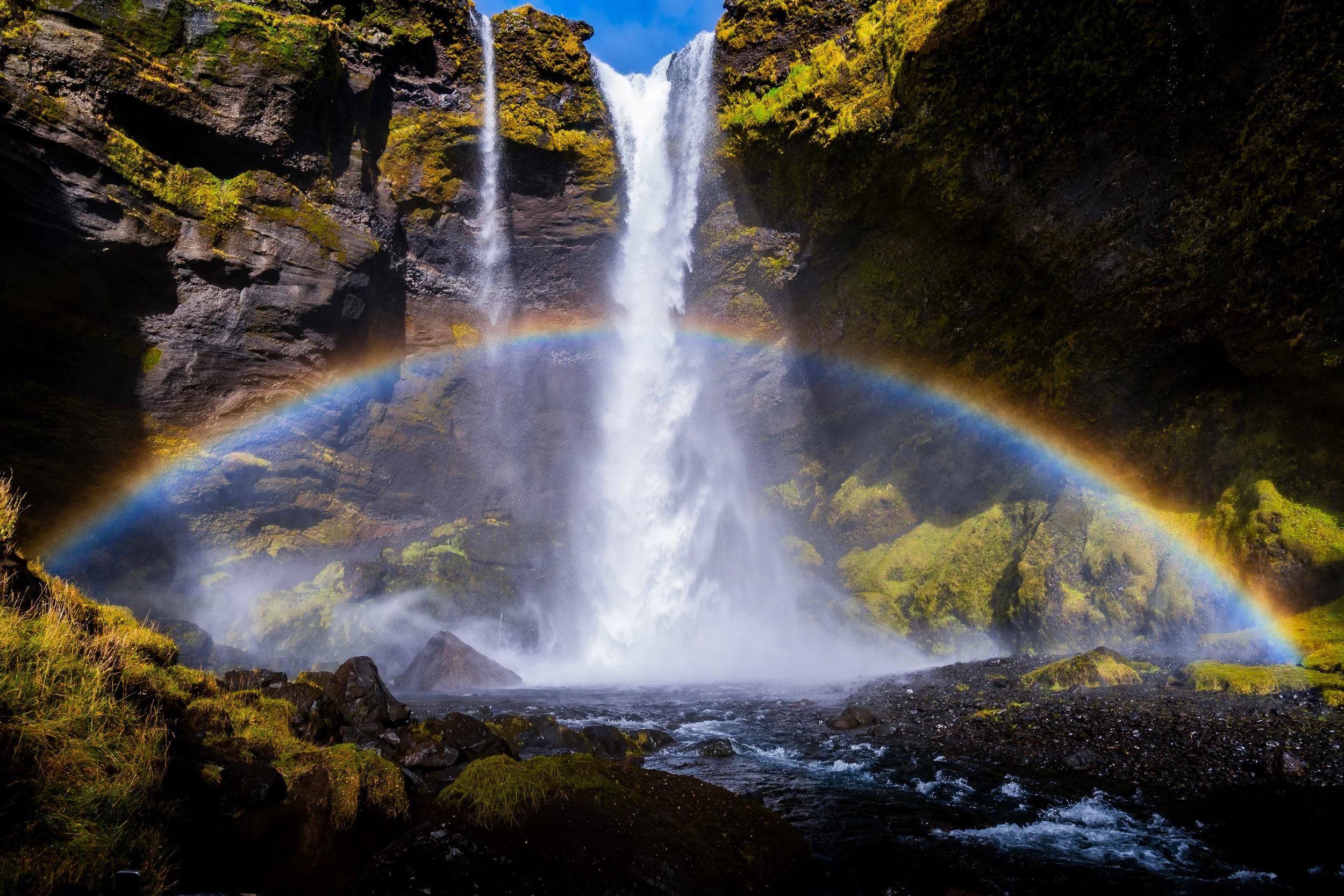 Waterfall cascading through a moss-covered canyon with a vivid rainbow in the mist