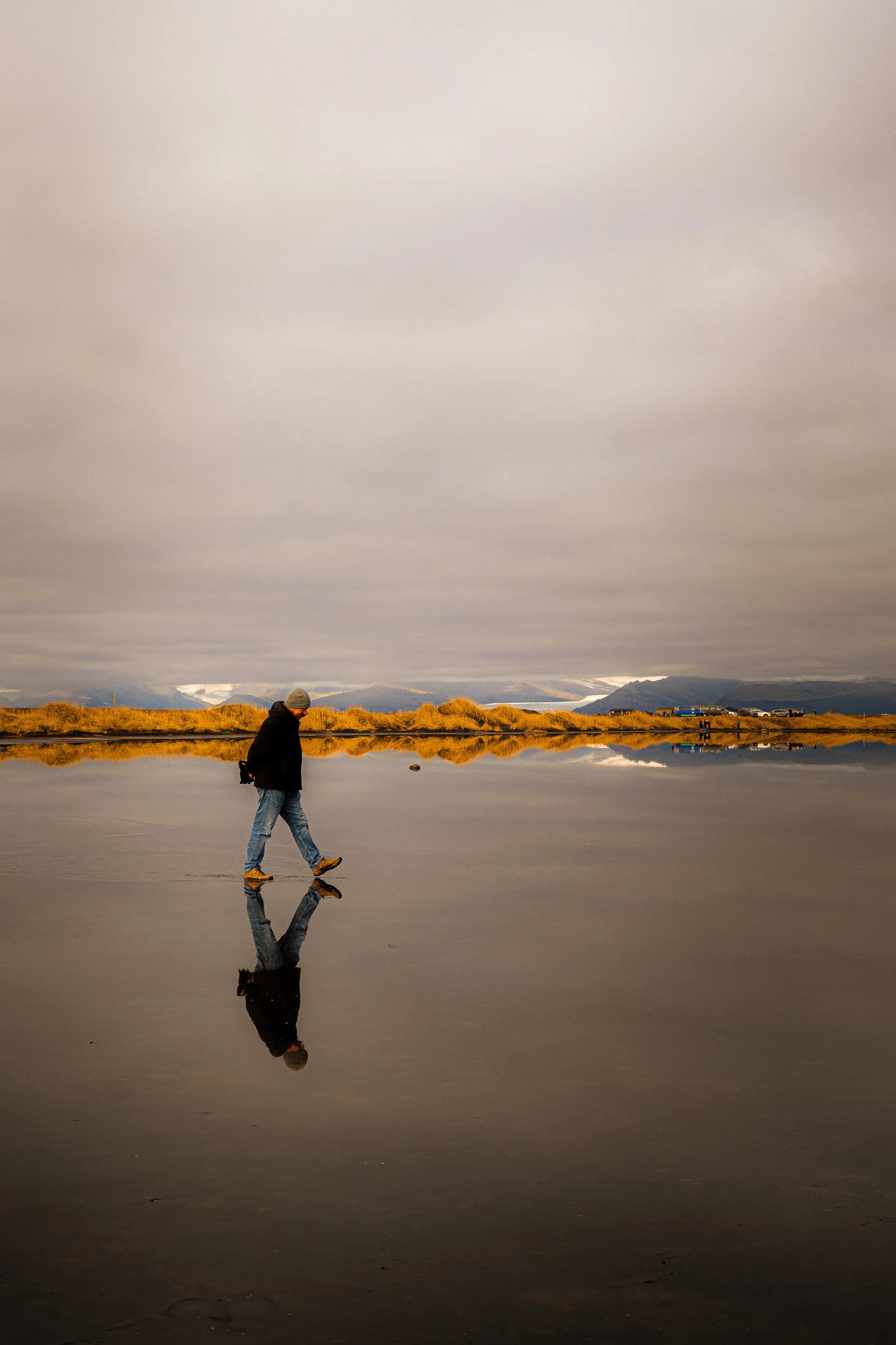 Person walking across reflective water at sunset with mountains and golden grasses in the background