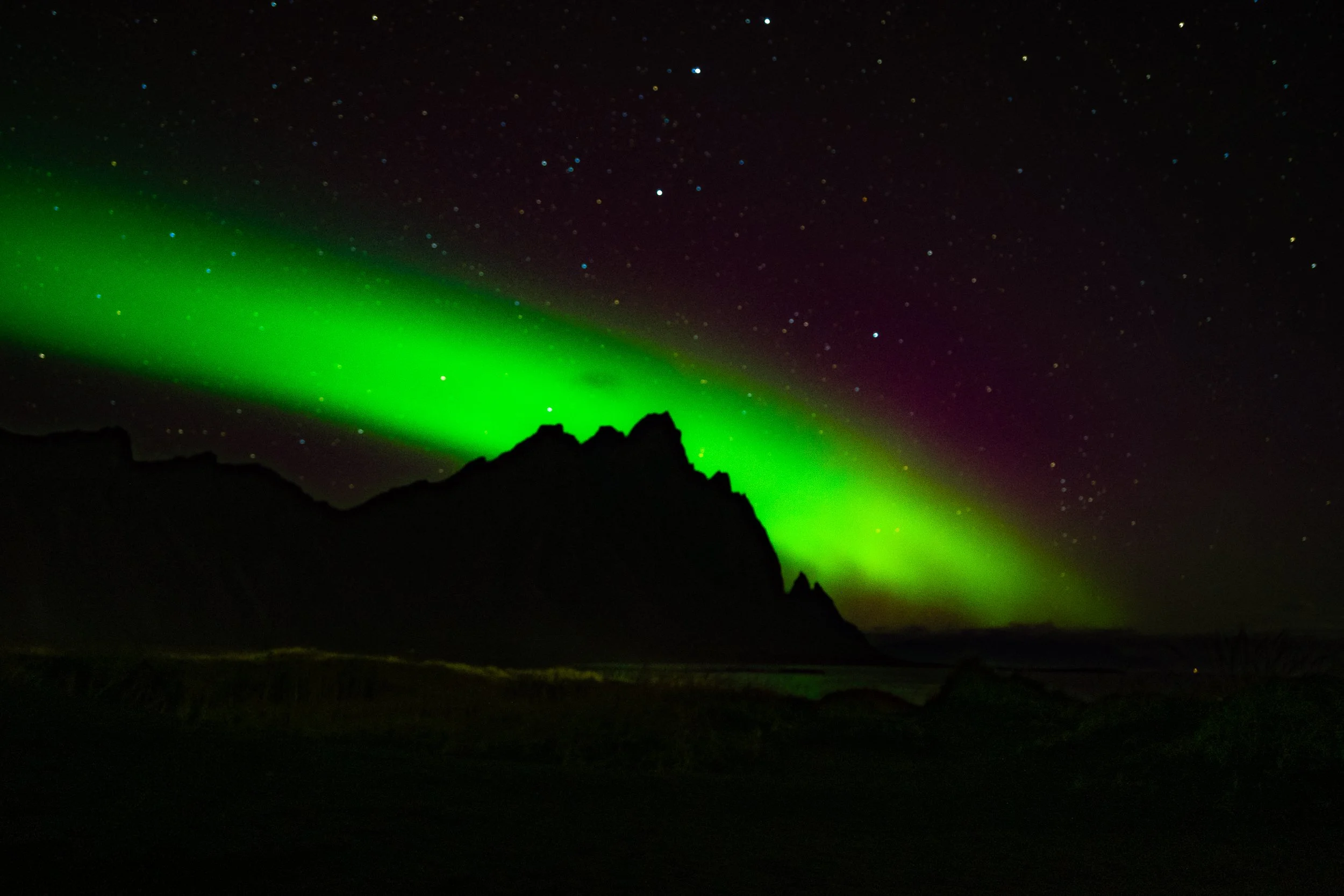 Bright green aurora borealis over a jagged mountain silhouette beneath a star-filled night sky