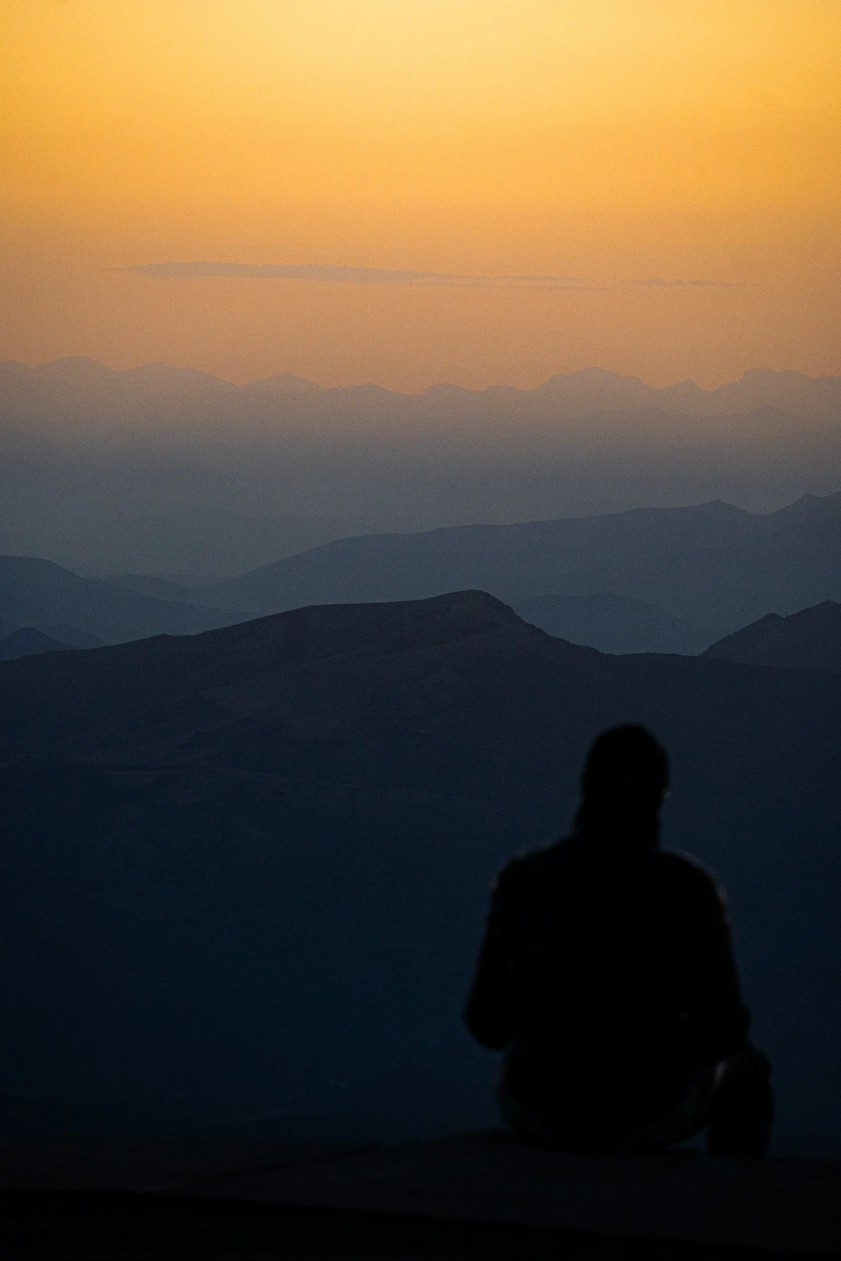 Silhouetted person sitting before layered mountain ranges at sunset