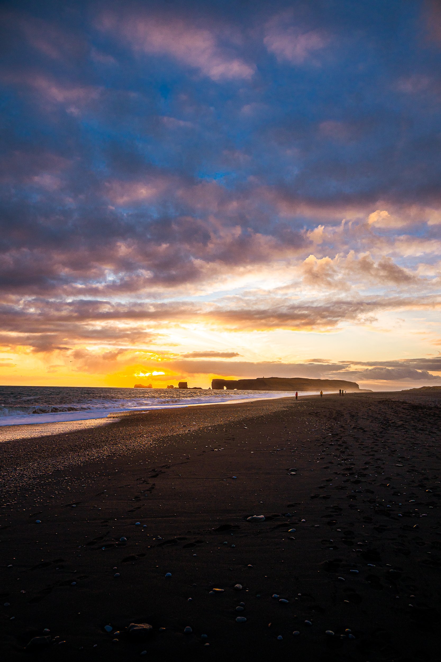 Sunset over a black sand beach with ocean waves, sea stacks, dramatic clouds, and distant beachgoers