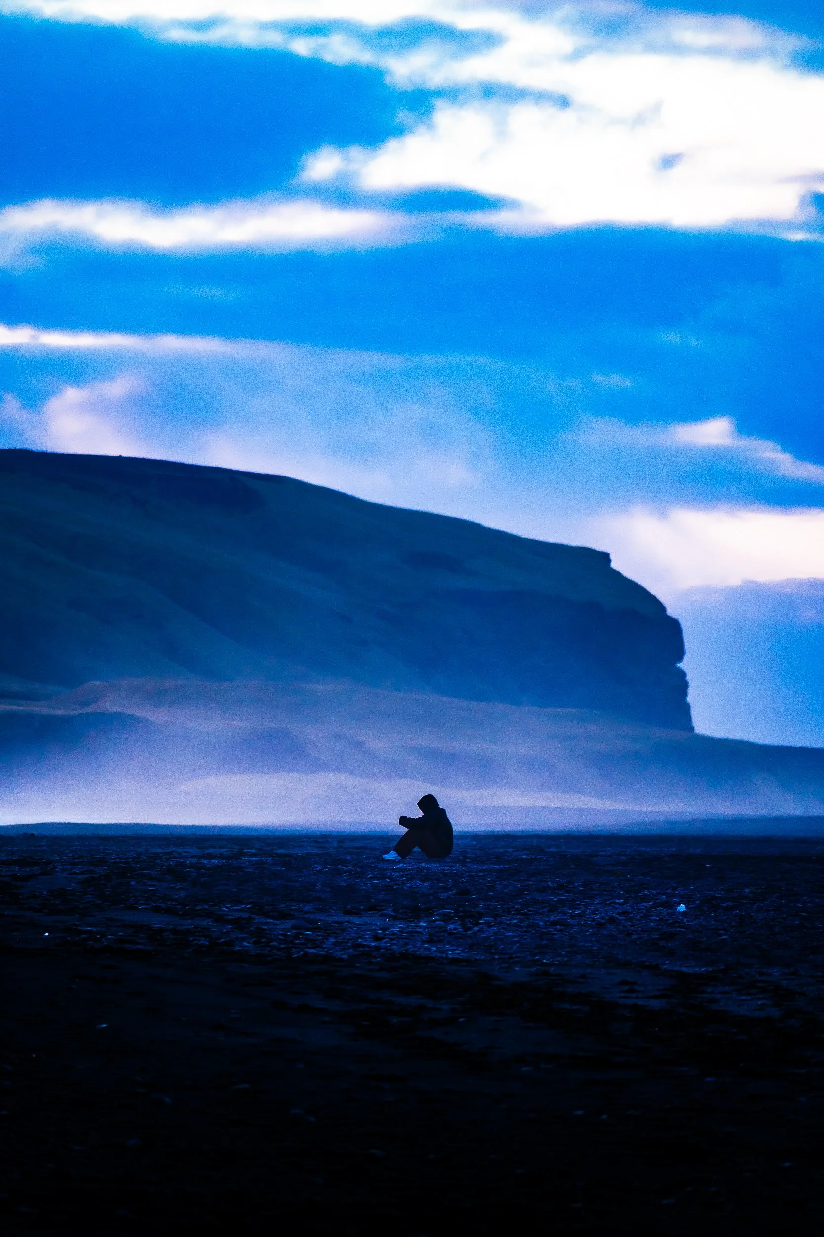 Silhouetted person sitting on a rocky shoreline beneath dramatic blue clouds and a dark coastal cliff