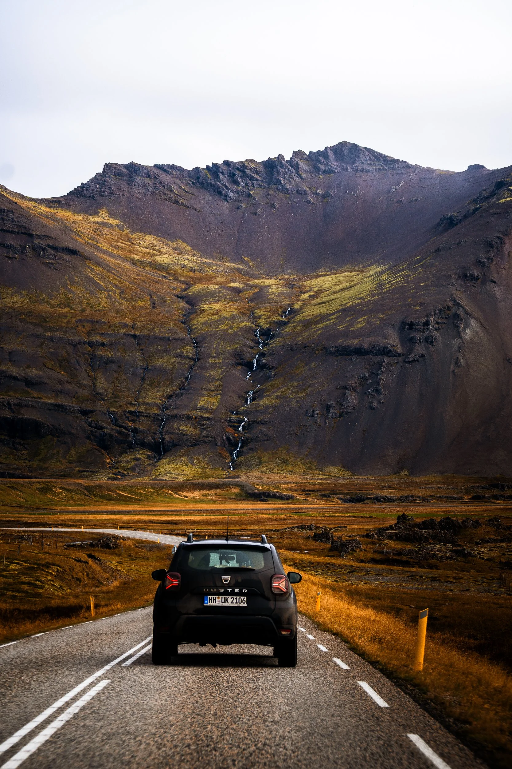 Dacia Duster driving on an Icelandic road beneath a rugged, moss-covered mountain with cascading streams