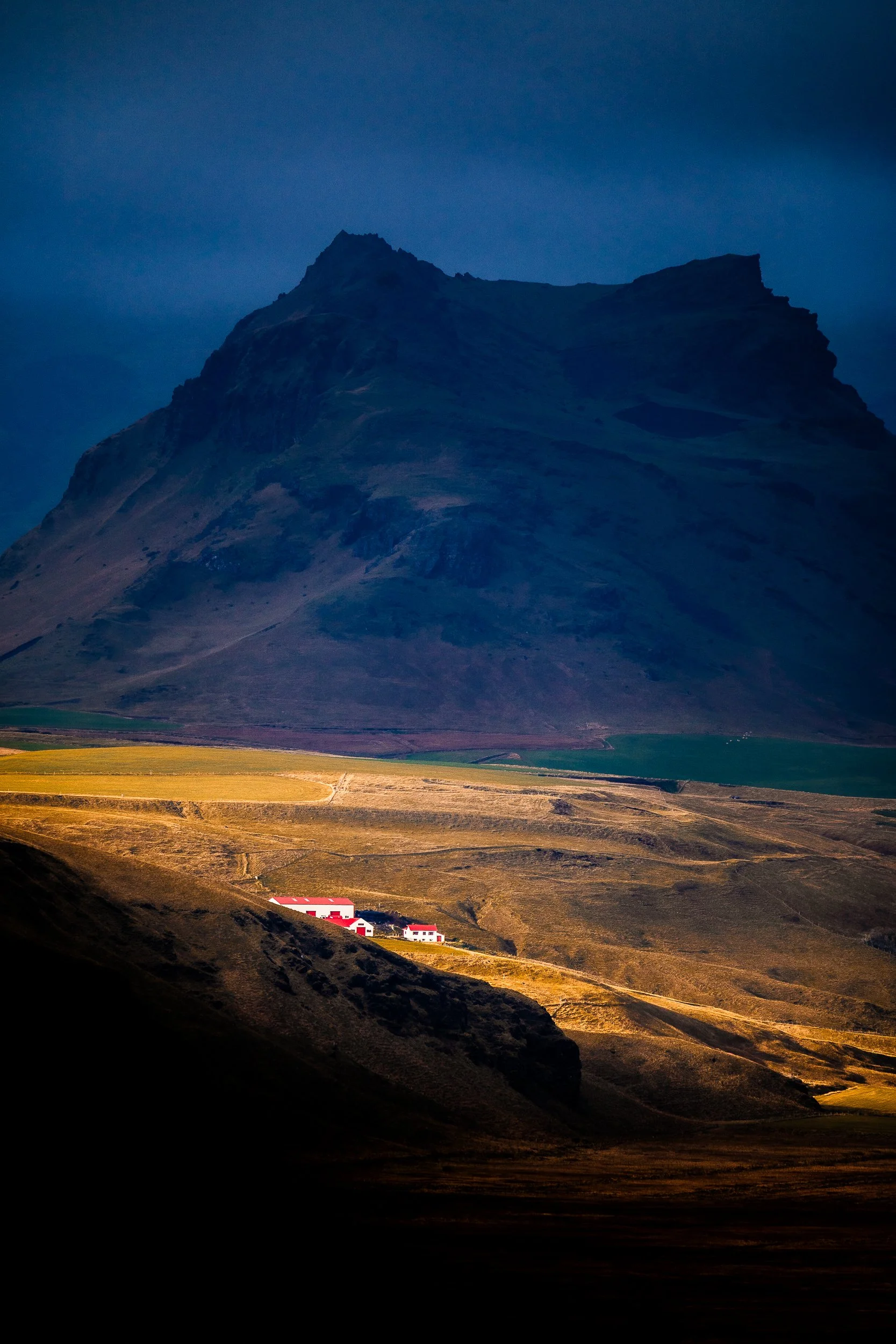 Dramatic mountain landscape with a rugged peak, colorful farmland, and small red-roofed buildings at sunset