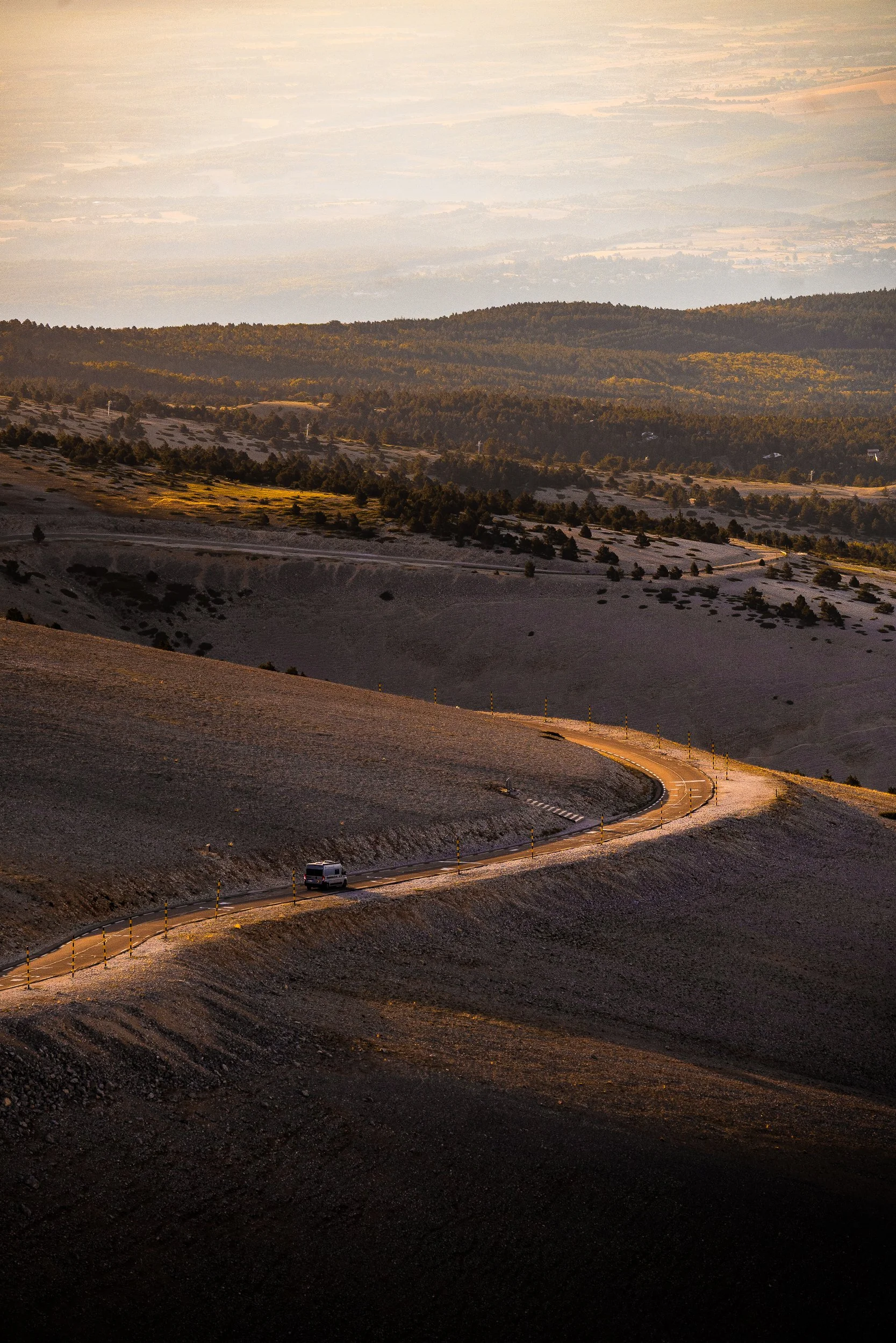 Winding mountain road with a van crossing a rocky, snow-dusted volcanic landscape above forested hills and a hazy valley at sunset