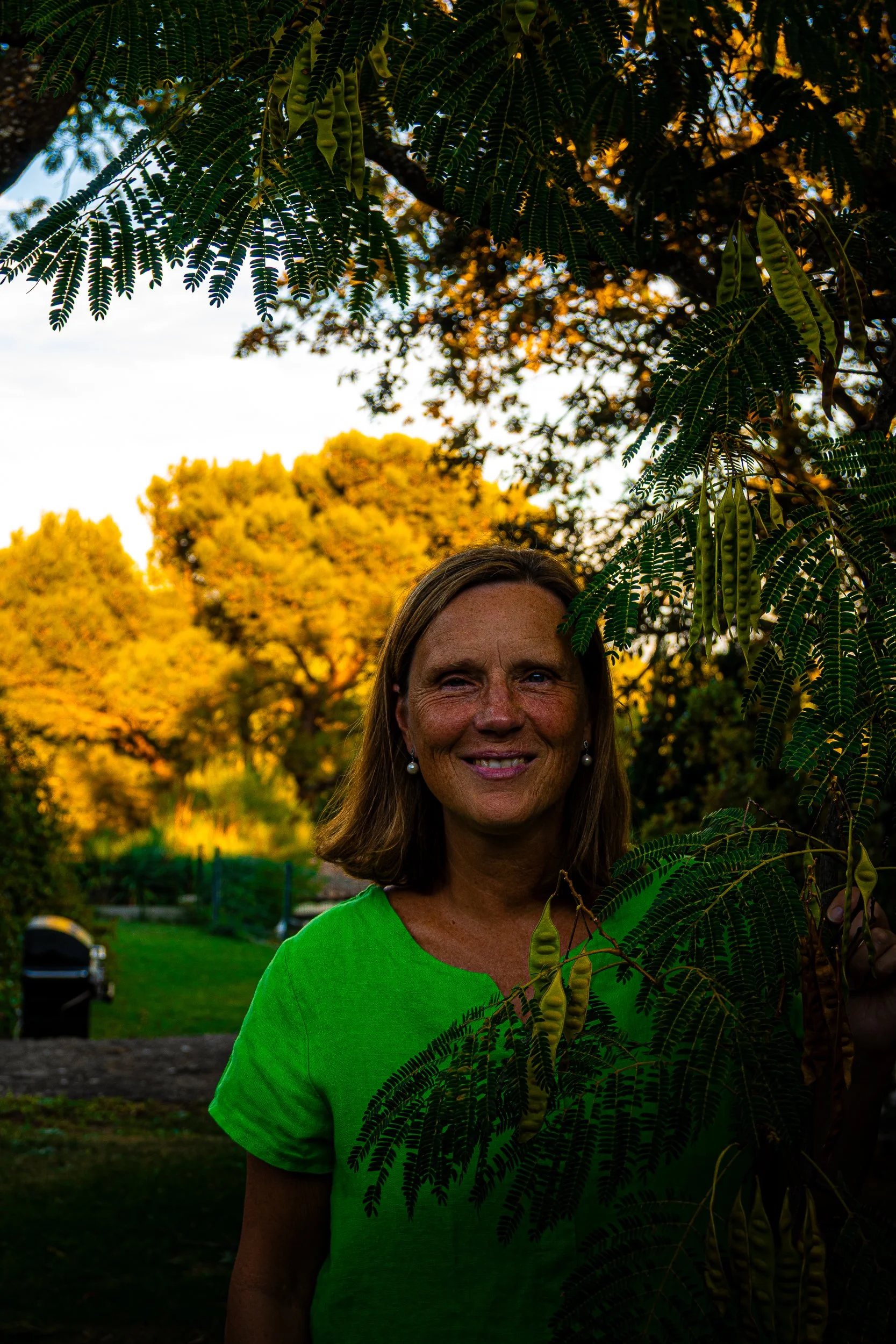 Smiling woman in a bright green shirt standing among feathery tree branches and seed pods in a sunlit garden