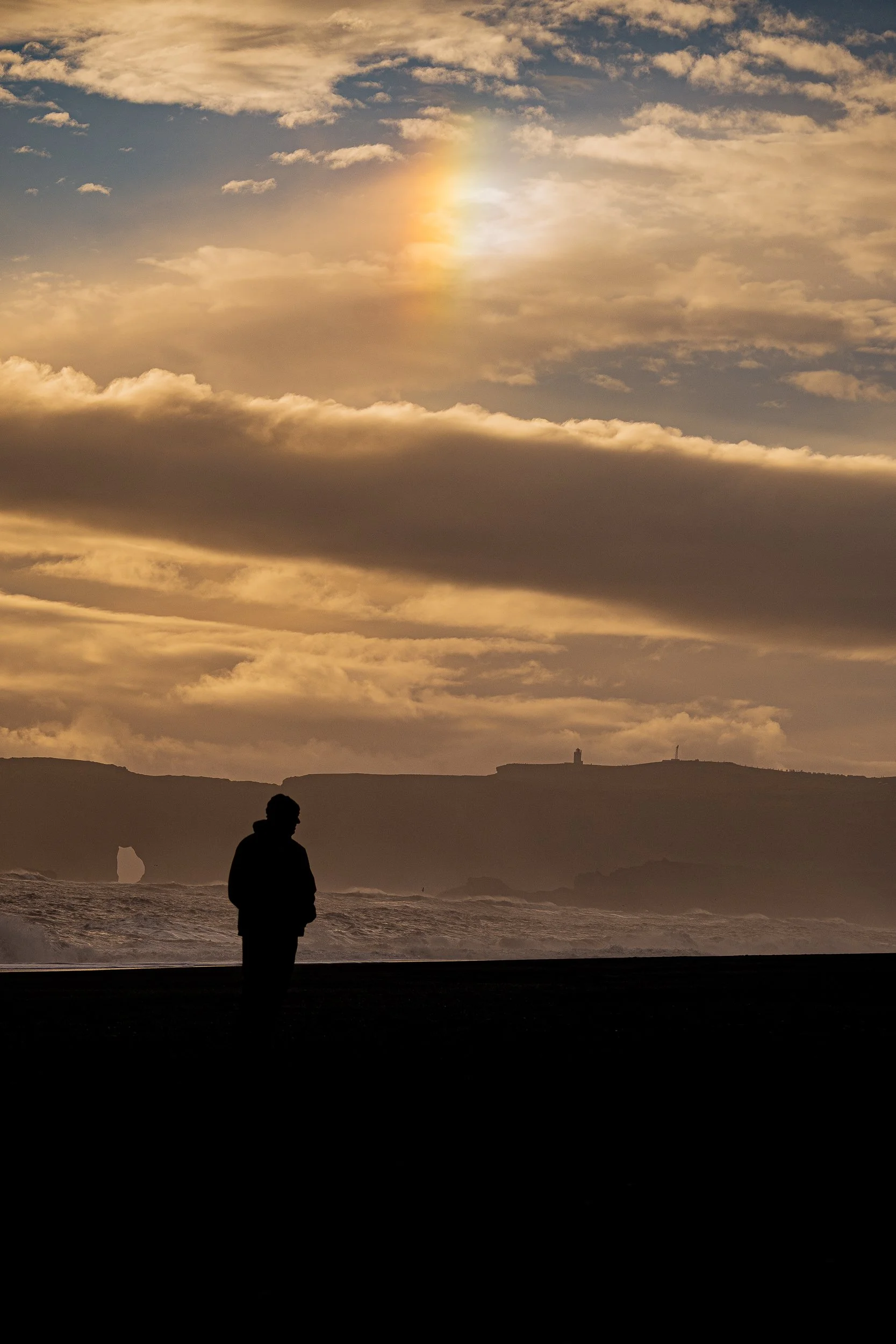 Silhouetted person standing beside a choppy ocean beneath a dramatic golden sunset and layered clouds, with a rocky coastline and lighthouse on the horizon.