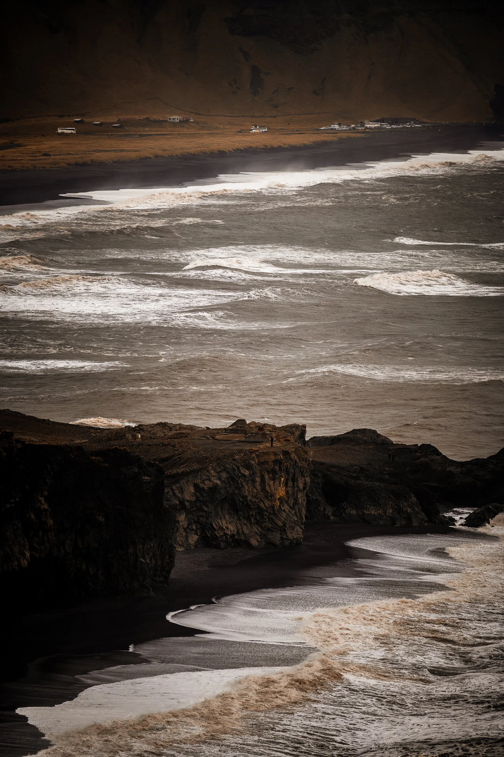 Powerful waves breaking along a black-sand beach beneath rugged volcanic cliffs, with tour buses and vehicles in the distance.