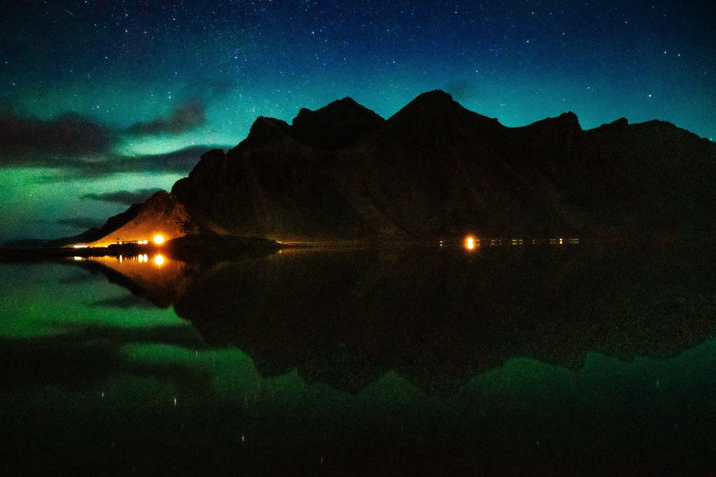Northern lights glowing over a starry night sky, silhouetted mountain peaks, and a dark lake with reflected lights